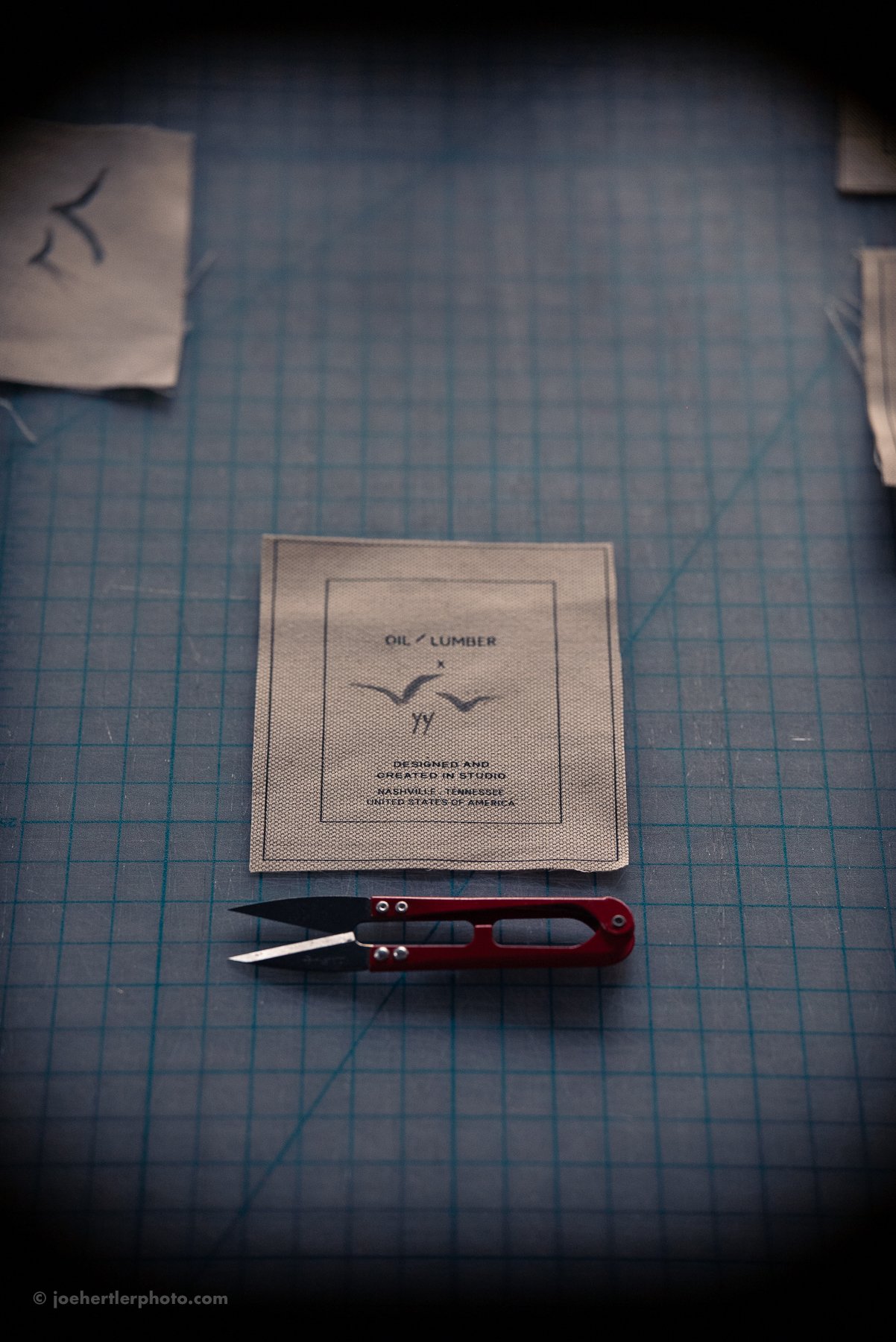 A pair of small scissors with a red handle, a fabric piece with printed text, and other fabric pieces on a blue cutting mat with grid lines.