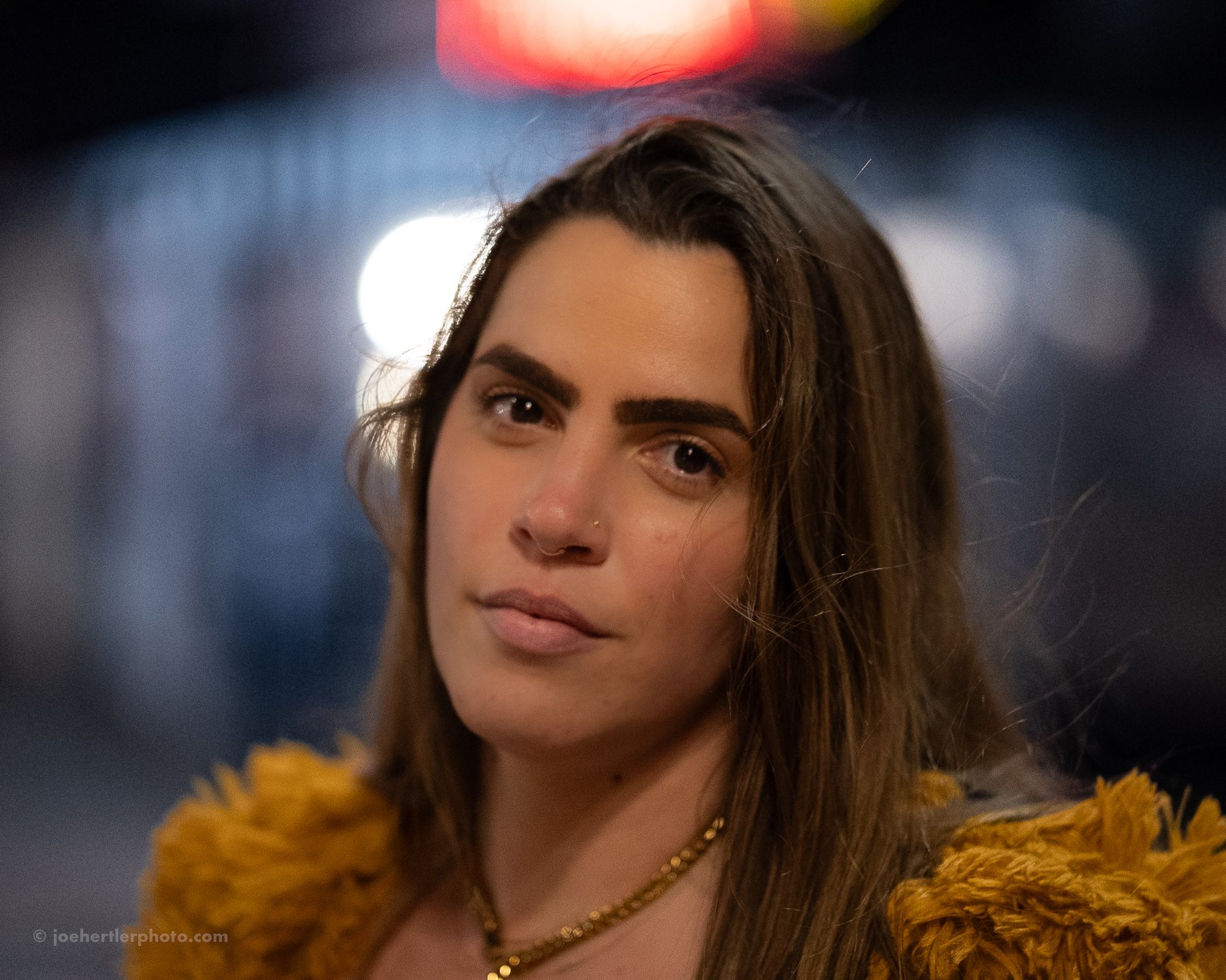 A woman with long brown hair, wearing a golden necklace and a rust-colored fuzzy jacket, standing outdoors at night with blurred city lights in the background.