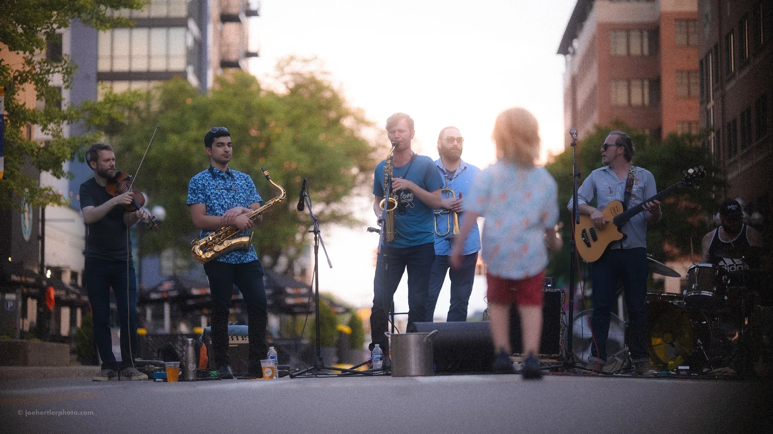 A street band of six musicians performing outdoors at sunset, with a woman in the foreground watching.