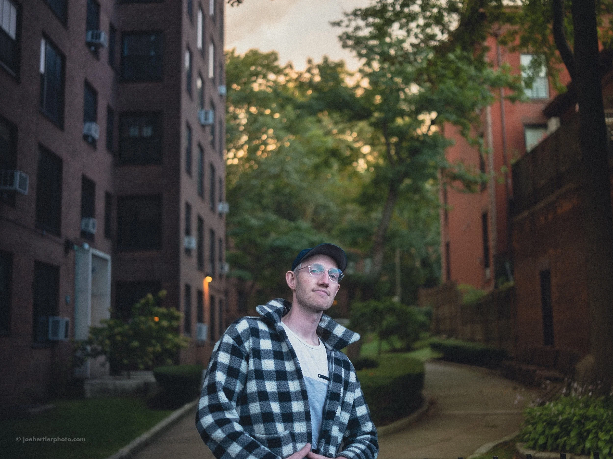 A young man with glasses wearing a black and white checkered jacket and a baseball cap stands outdoors on a path between brown brick apartment buildings with trees and bushes during sunset.