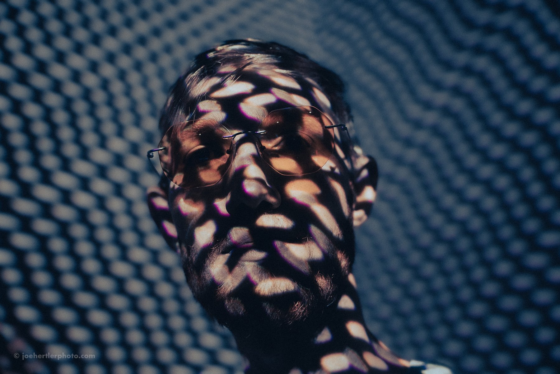 A man with a beard and glasses is illuminated by light projecting an abstract pattern of pink, purple, and orange shapes onto his face and background.