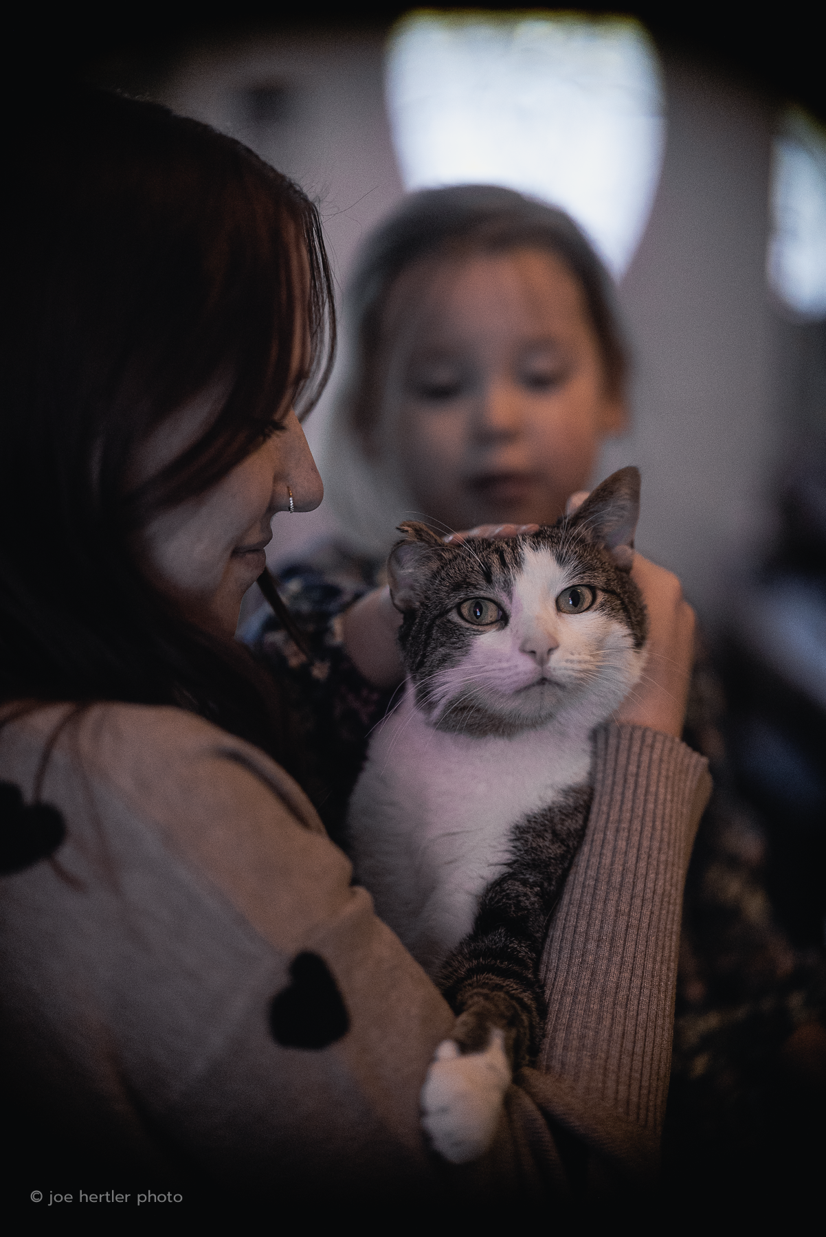 A woman holding a tabby cat with white fur on its face and chest, while a young girl looks at the cat in the background.