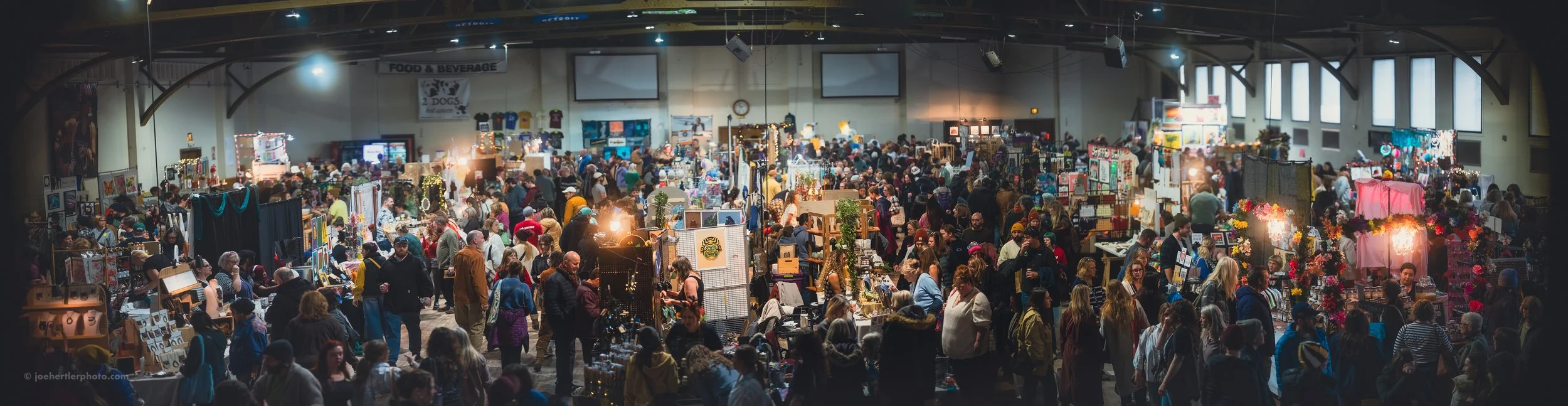 A crowded indoor market with numerous vendors and shoppers, colorful items and displays, and high ceilings with windows.