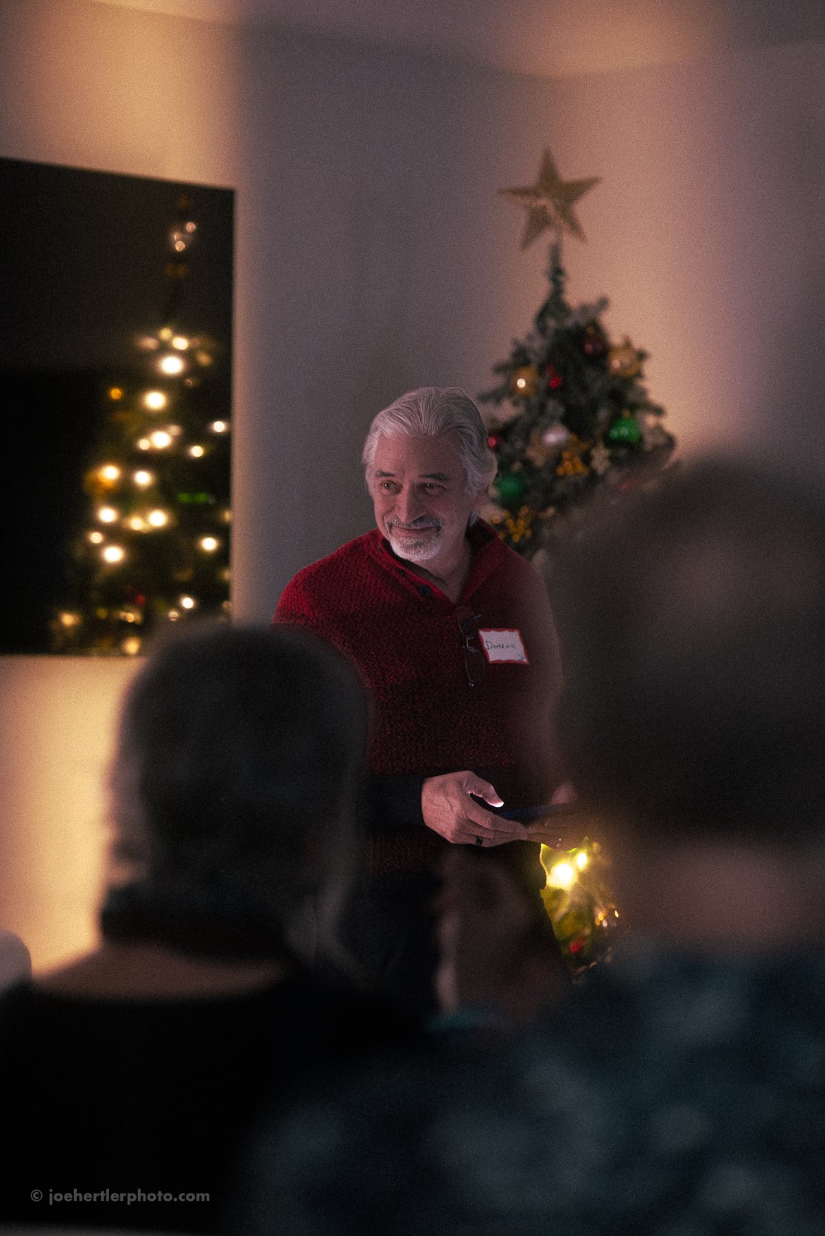 A man with gray hair and a beard, wearing a red sweater, stands in a dimly lit room decorated with Christmas trees and ornaments, holding a phone and smiling.