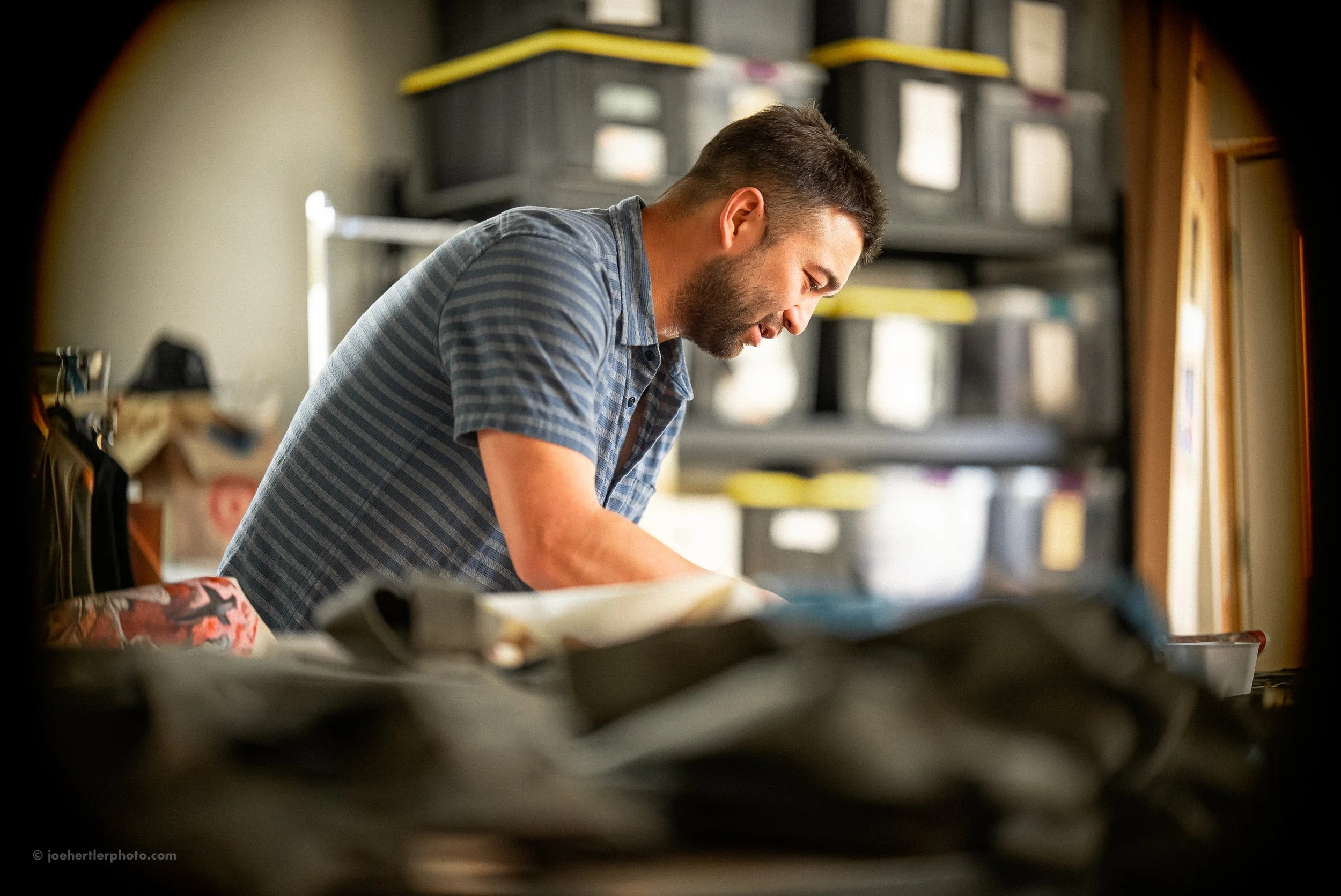 A man with dark hair and a beard, wearing a blue checkered shirt, leaning forward and working on a task in a room with storage bins and shelves in the background.