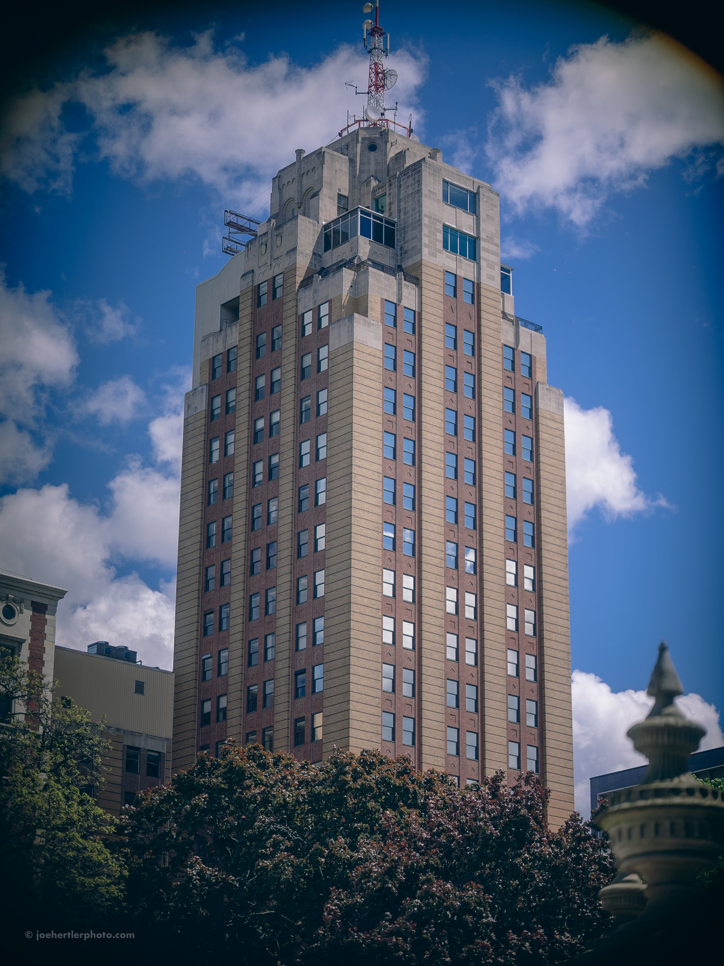 Tall skyscraper with modern architectural design, topped with antennas and communication equipment, against a partly cloudy blue sky. Trees are in the foreground.