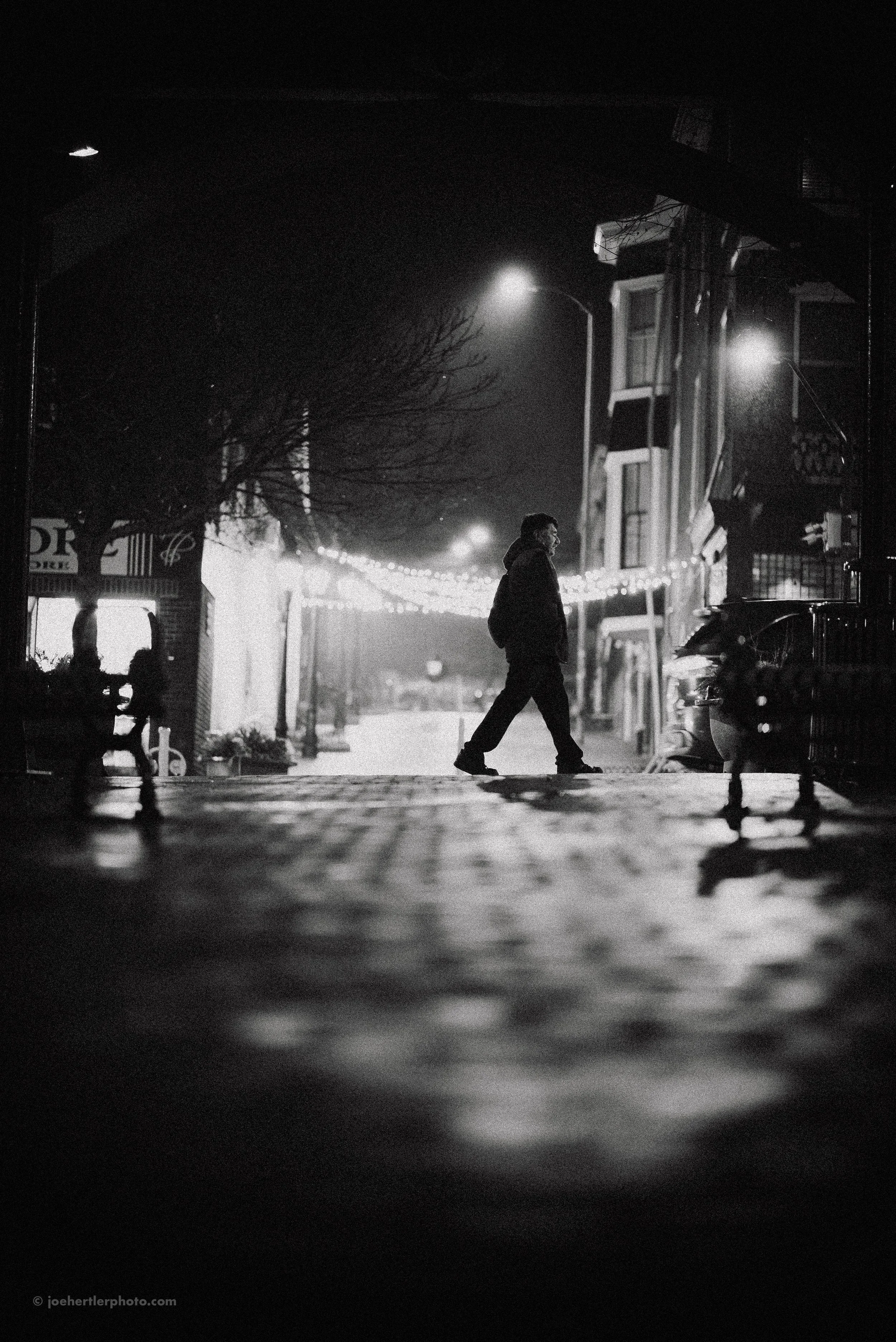 A man walking alone on a city street at night, with streetlights and buildings in the background, in black and white.