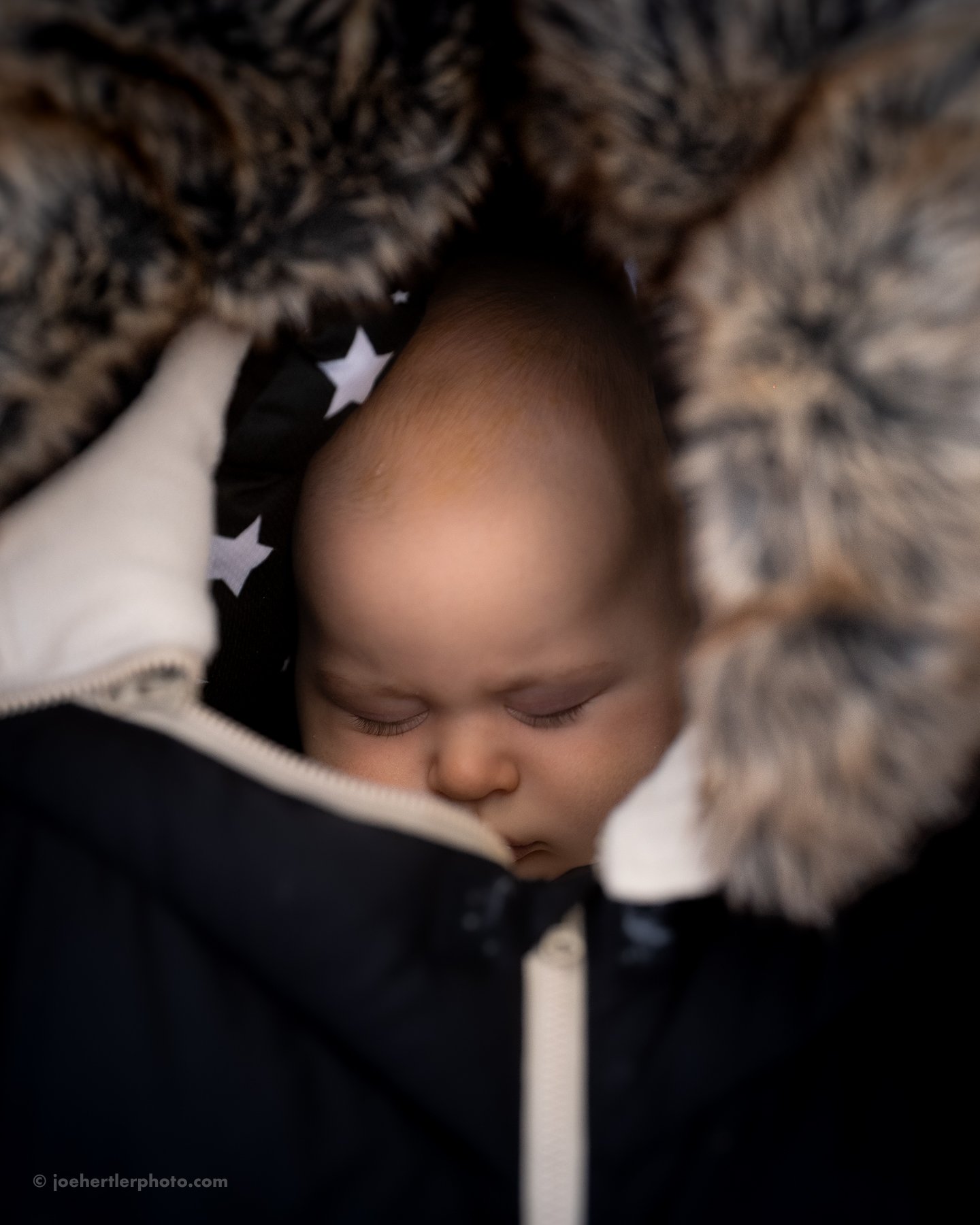 Close-up of a sleeping baby with closed eyes, wearing a black jacket with a fur-lined hood, nestled in a cozy blanket with black and white stars.
