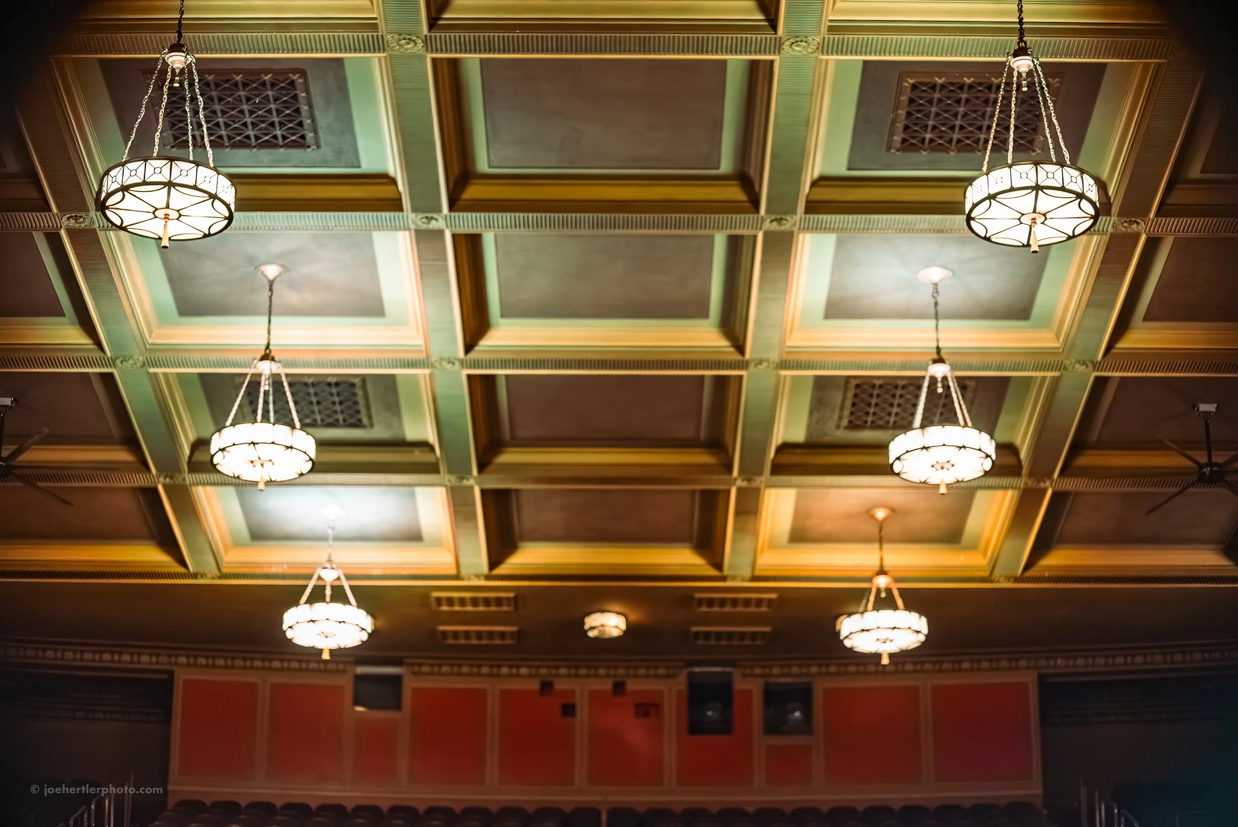 Photo of a decorated theater or banquet hall ceiling with ornate molding, green and brown textured panels, and six hanging chandelier lights.
