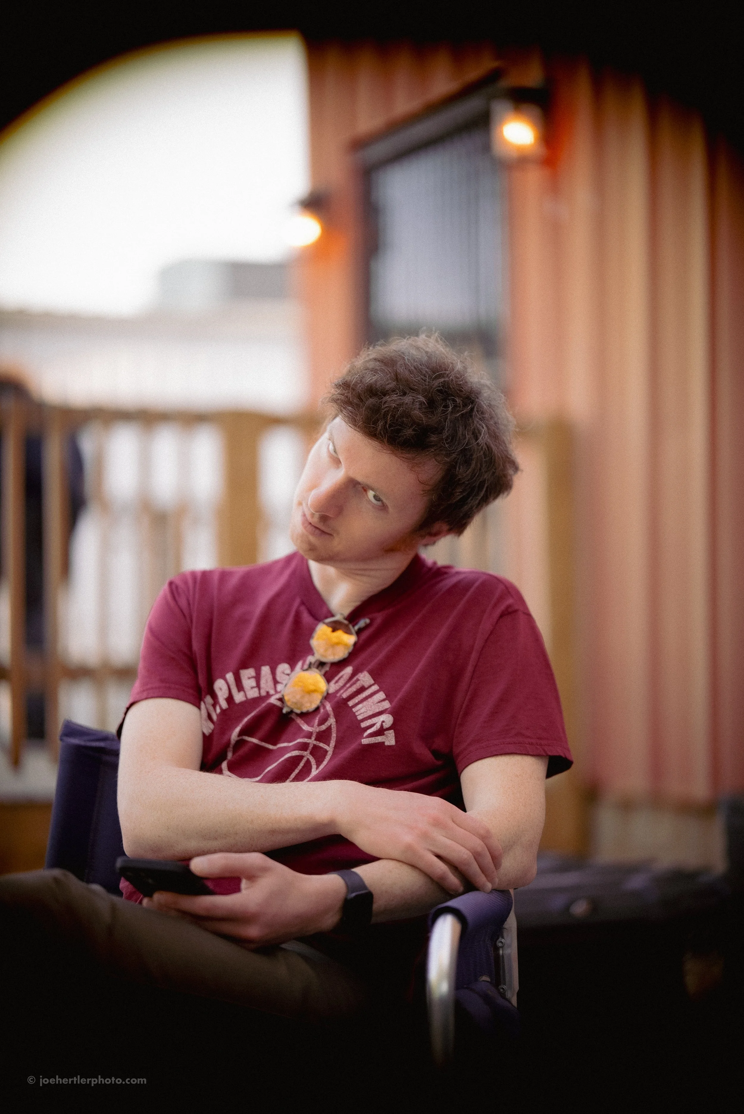A young man with curly dark hair wearing a maroon t-shirt sitting outdoors, looking up with his head tilted, holding a smartphone in his hand.