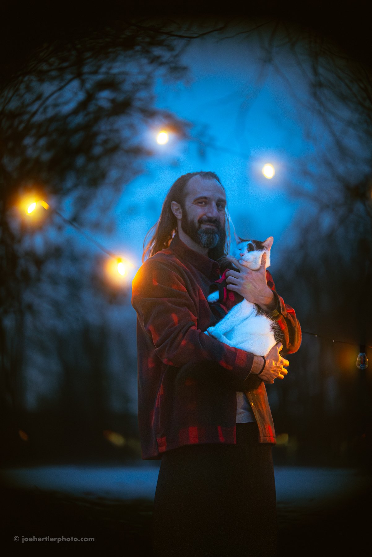 A man with long hair and a beard holding a white and black cat during twilight, with blue sky and string lights in the background.