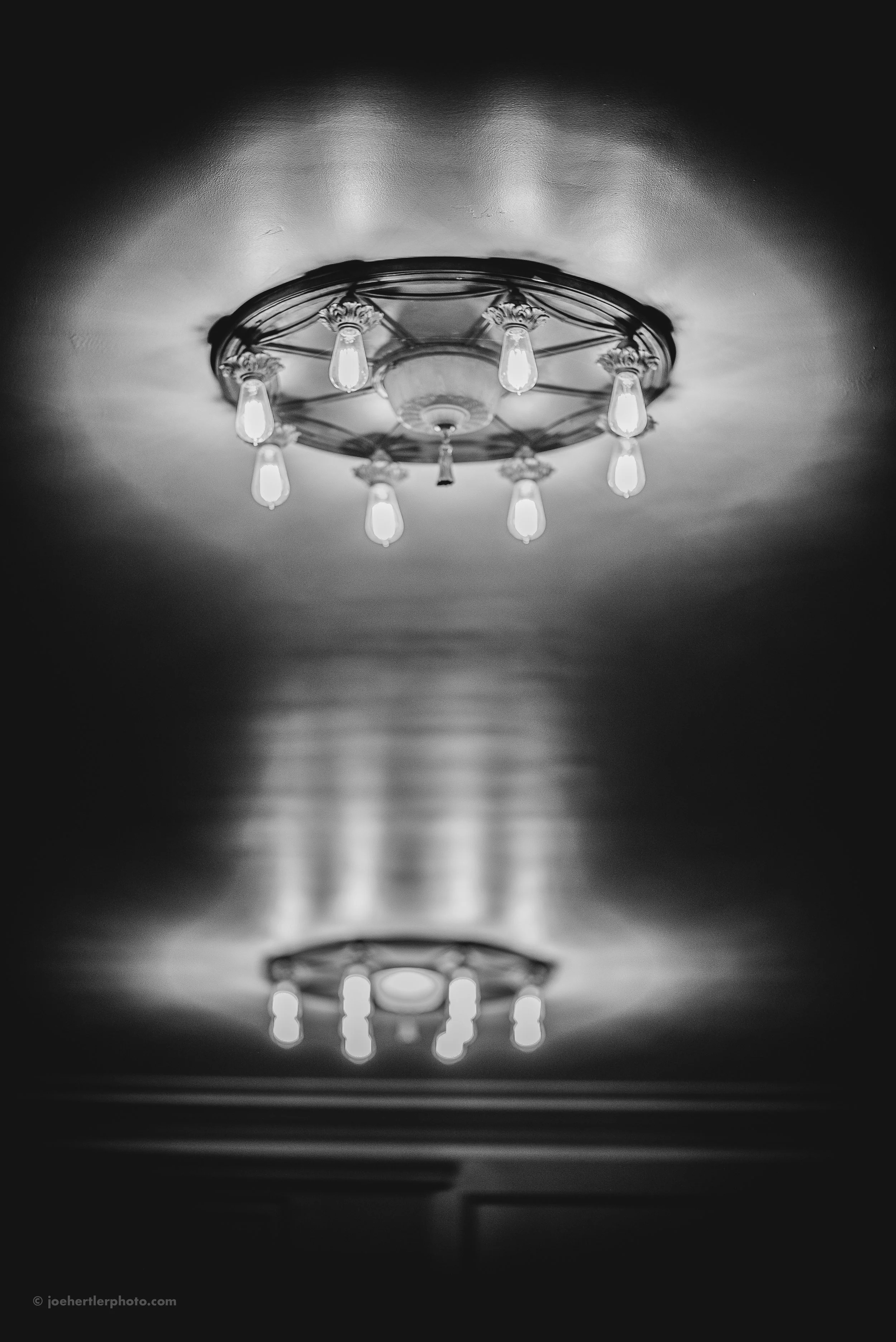 Black and white photo of a ceiling light fixture with multiple exposed bulbs and decorative elements, casting reflections and shadows on the ceiling.