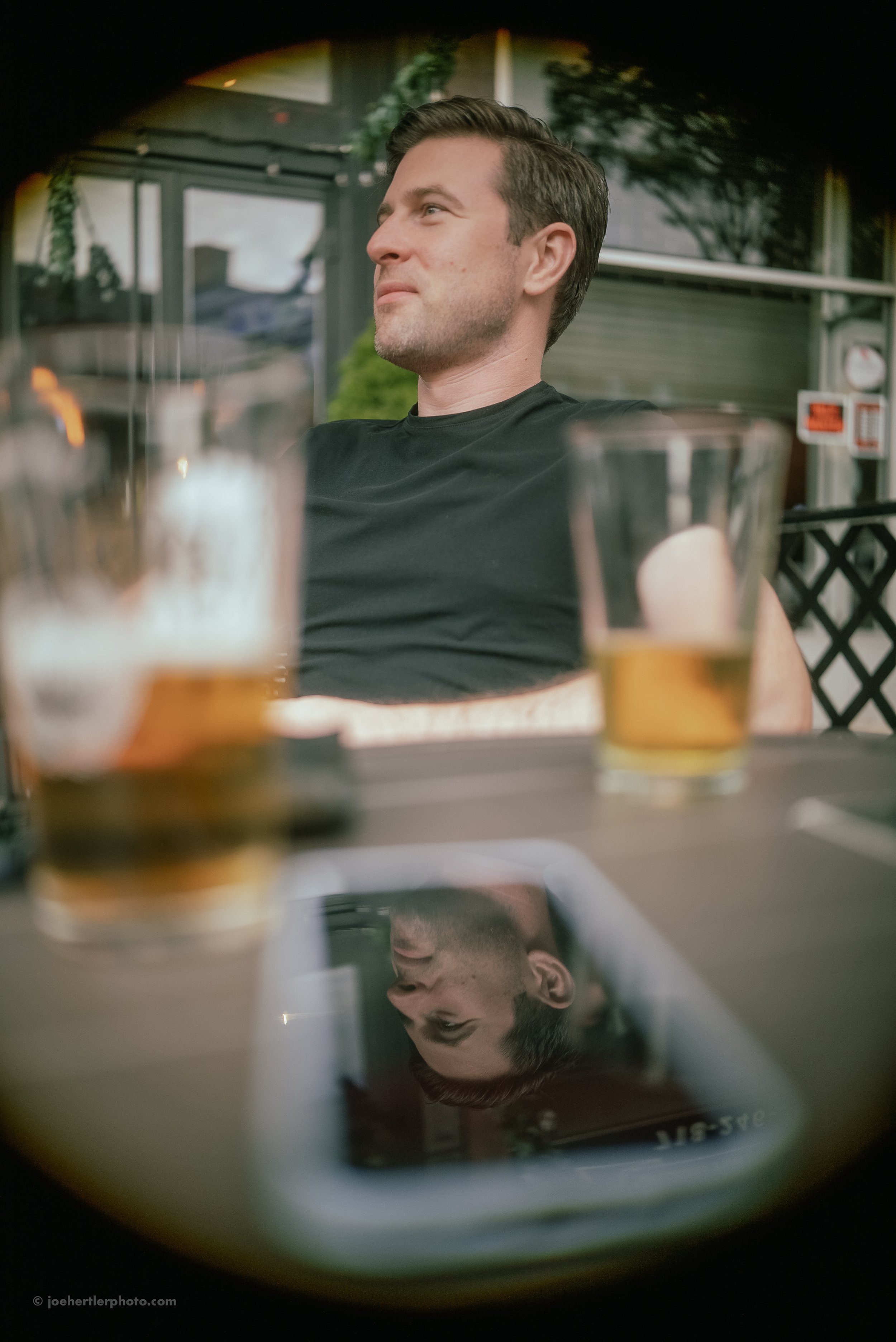 A man with short brown hair and a beard sits outdoors at a table, wearing a black t-shirt, with drinks in front of him, and his reflection is visible on a phone screen.