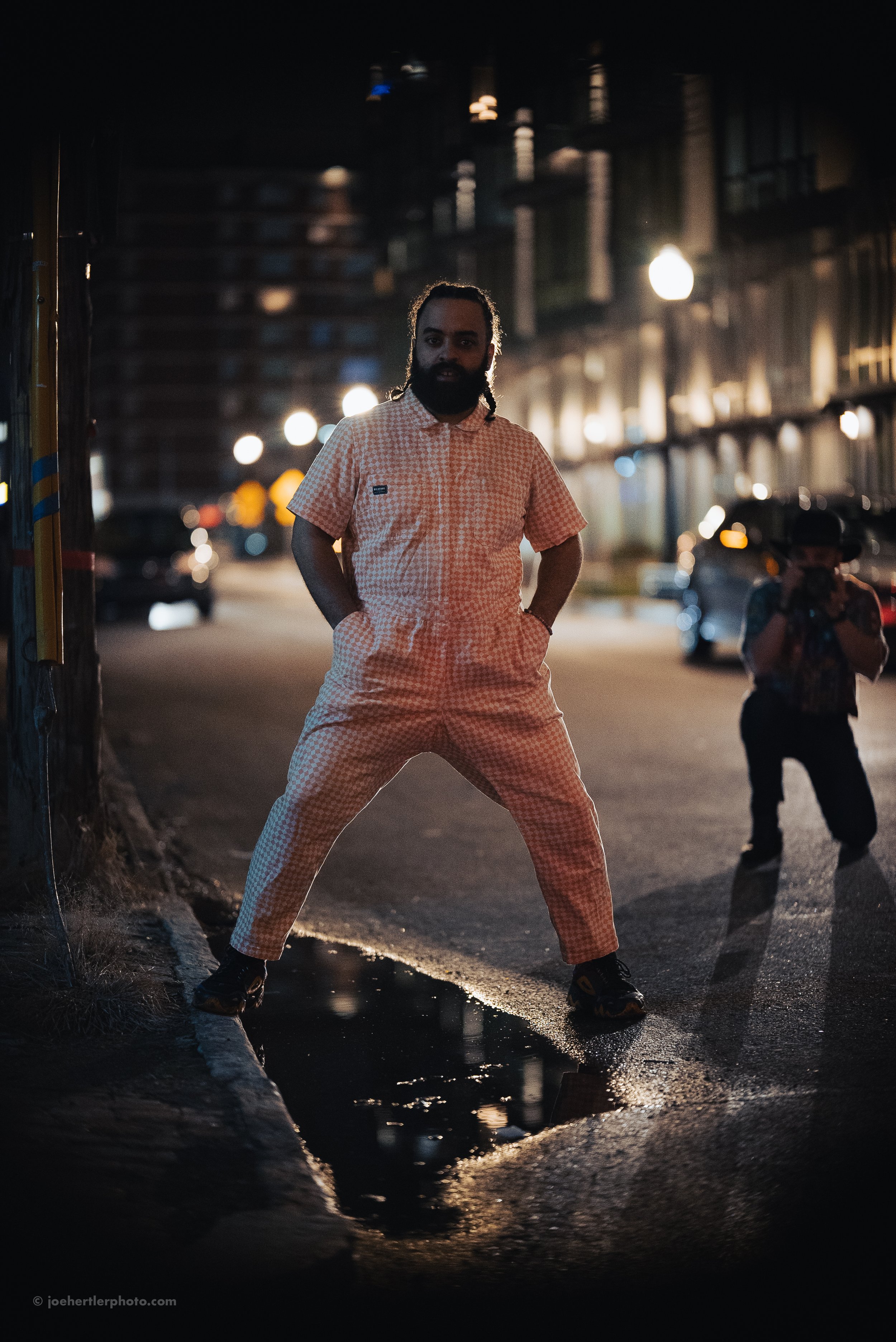 A man with a beard and long hair standing in a reflective puddle on a city street at night, with his hands in his pockets, wearing a checkered outfit. There are blurred city lights and buildings in the background, and another person taking photos is 