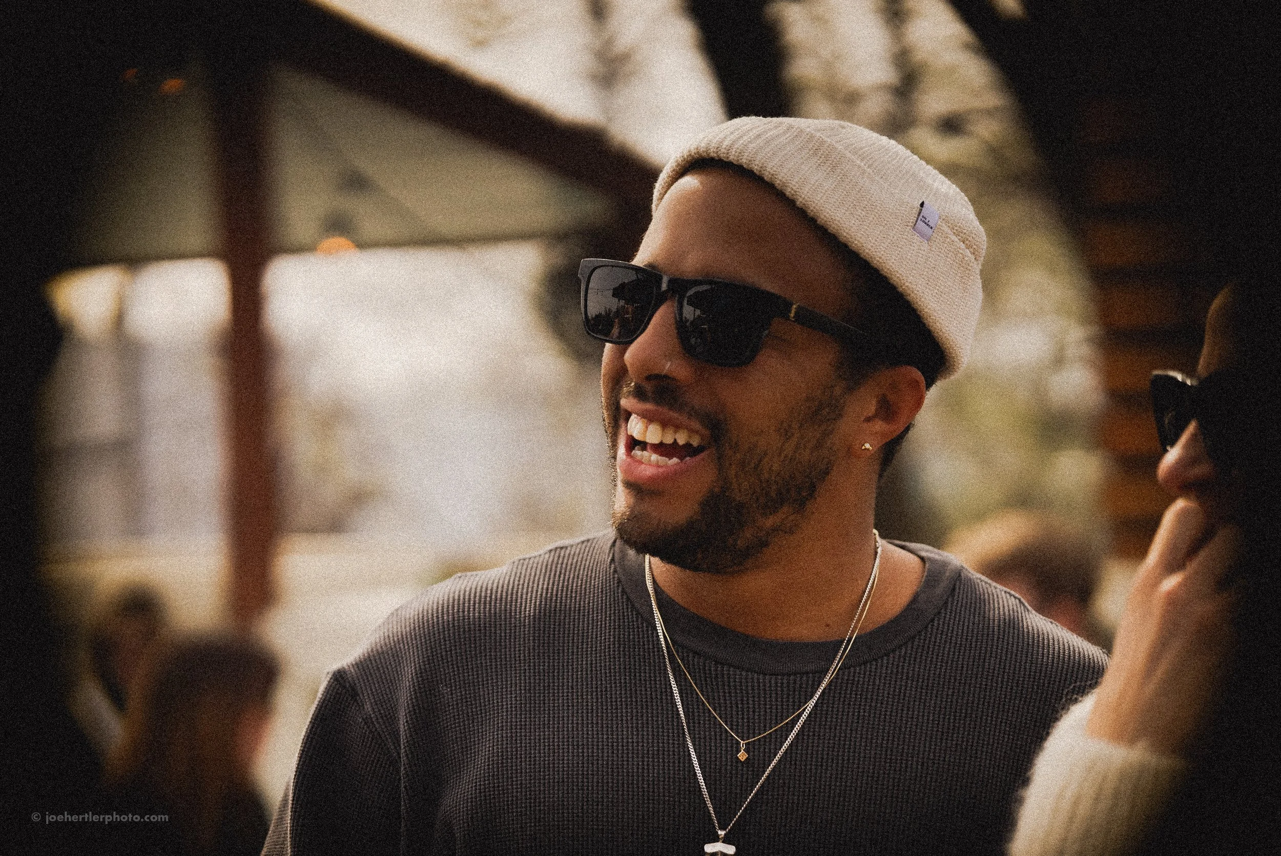A young man with a beard and dark skin, wearing black sunglasses, a beige beanie, and layered necklaces, is smiling and talking to someone.