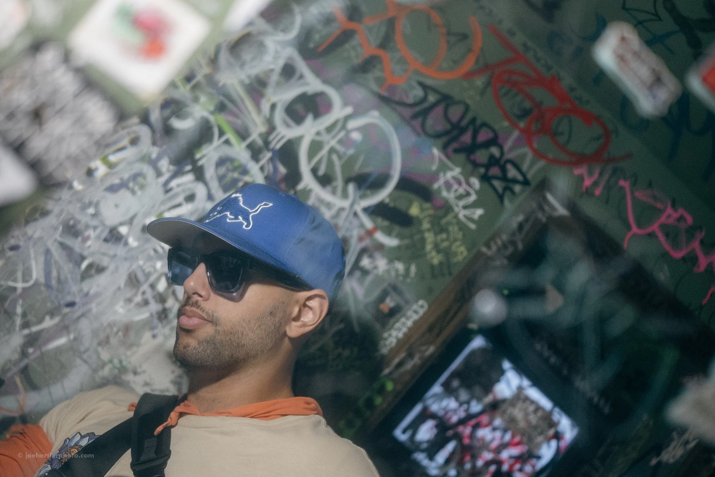 A man wearing a blue cap with a white logo and black sunglasses, leaning against a graffiti-covered wall.