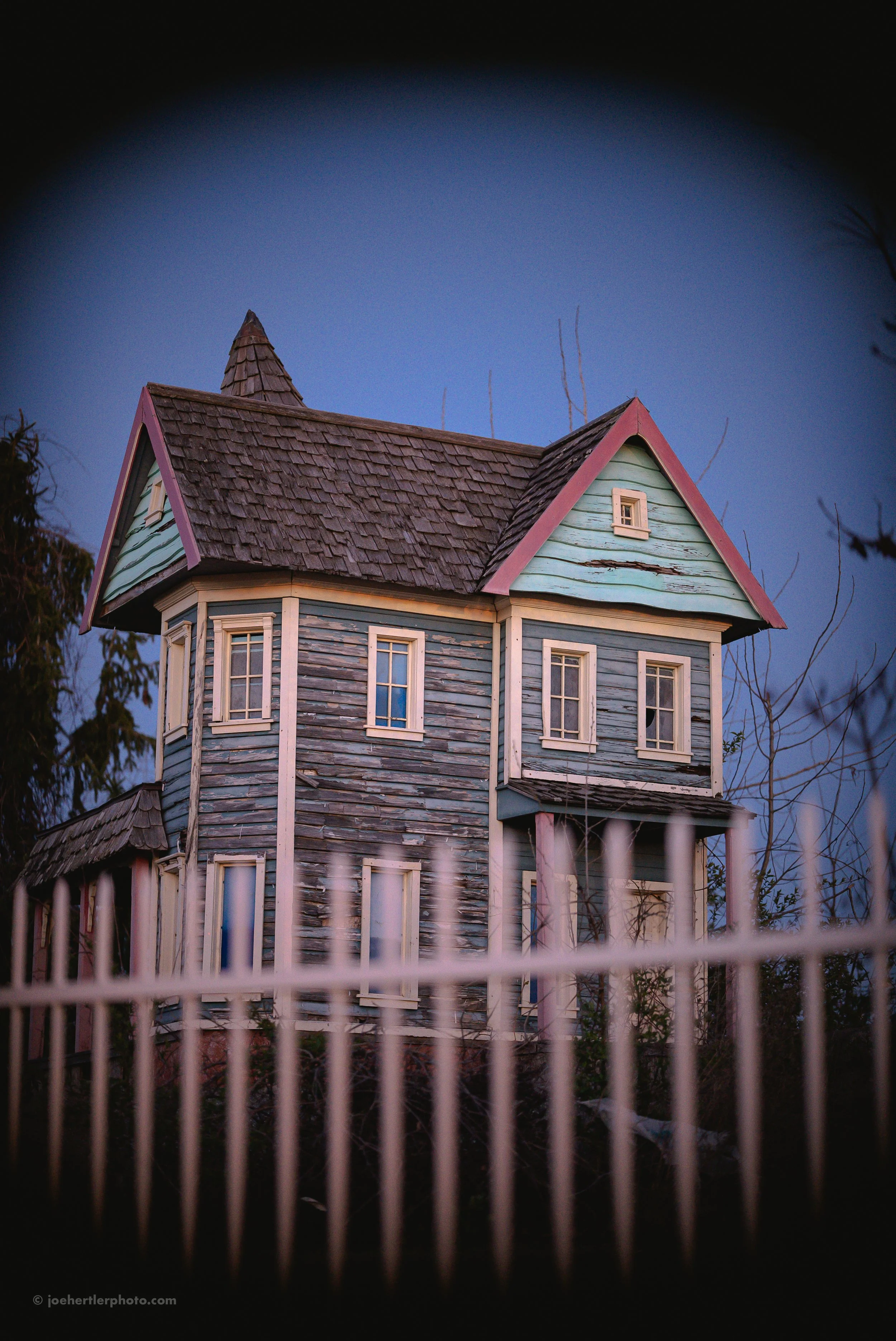 An old, weathered Victorian-style house with faded blue and mint green paint, surrounded by a white picket fence, captured during dusk.