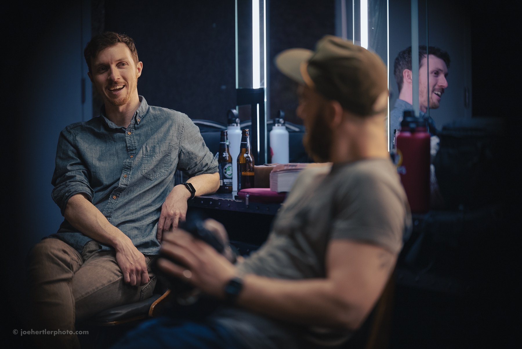 Two men having a conversation in a dimly lit bar or recording studio, one is sitting on a stool looking at the other, who is holding a gaming controller and wearing a cap, with beer bottles and other items on the counter behind them.
