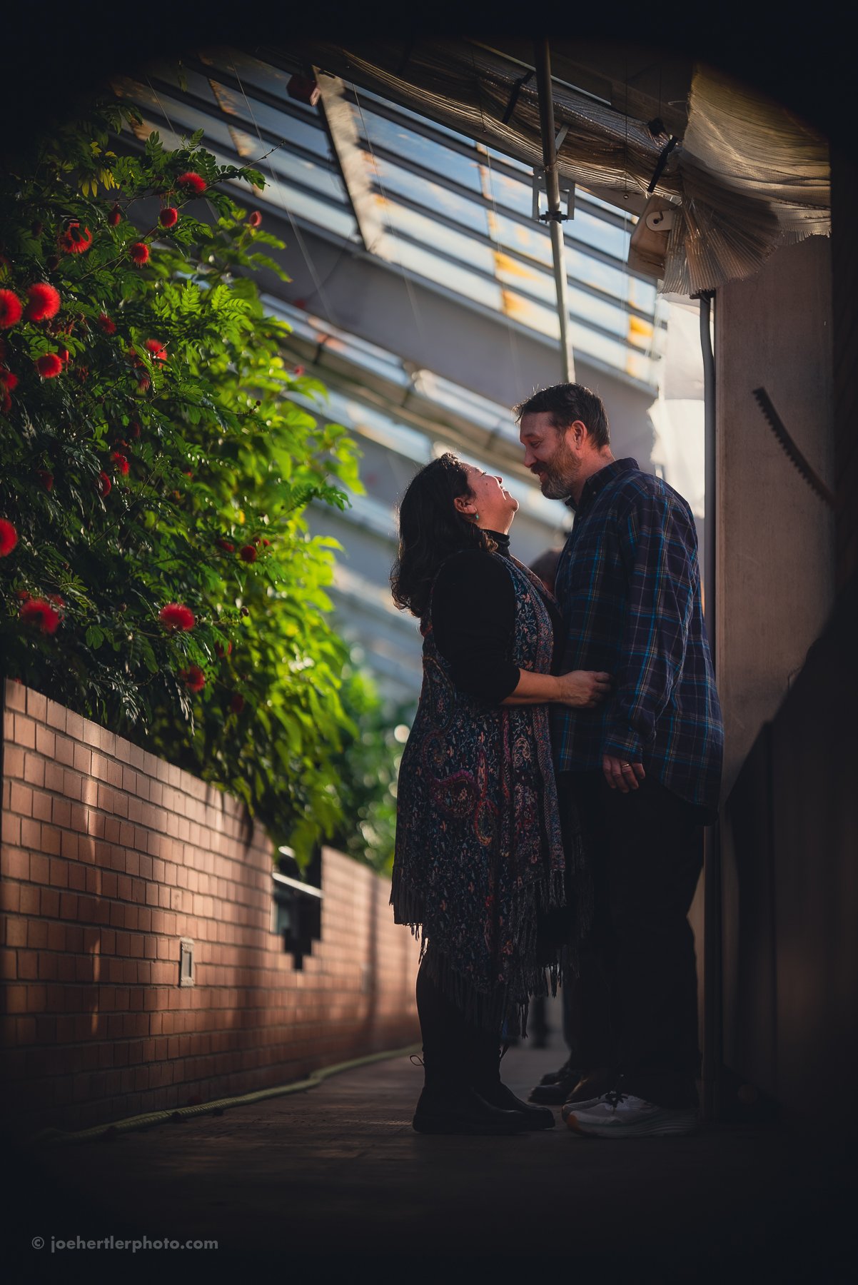 A couple stands close together, smiling and looking into each other's eyes, in an indoor garden or greenhouse with sunlight coming through a glass ceiling planted with green foliage and red flowers.