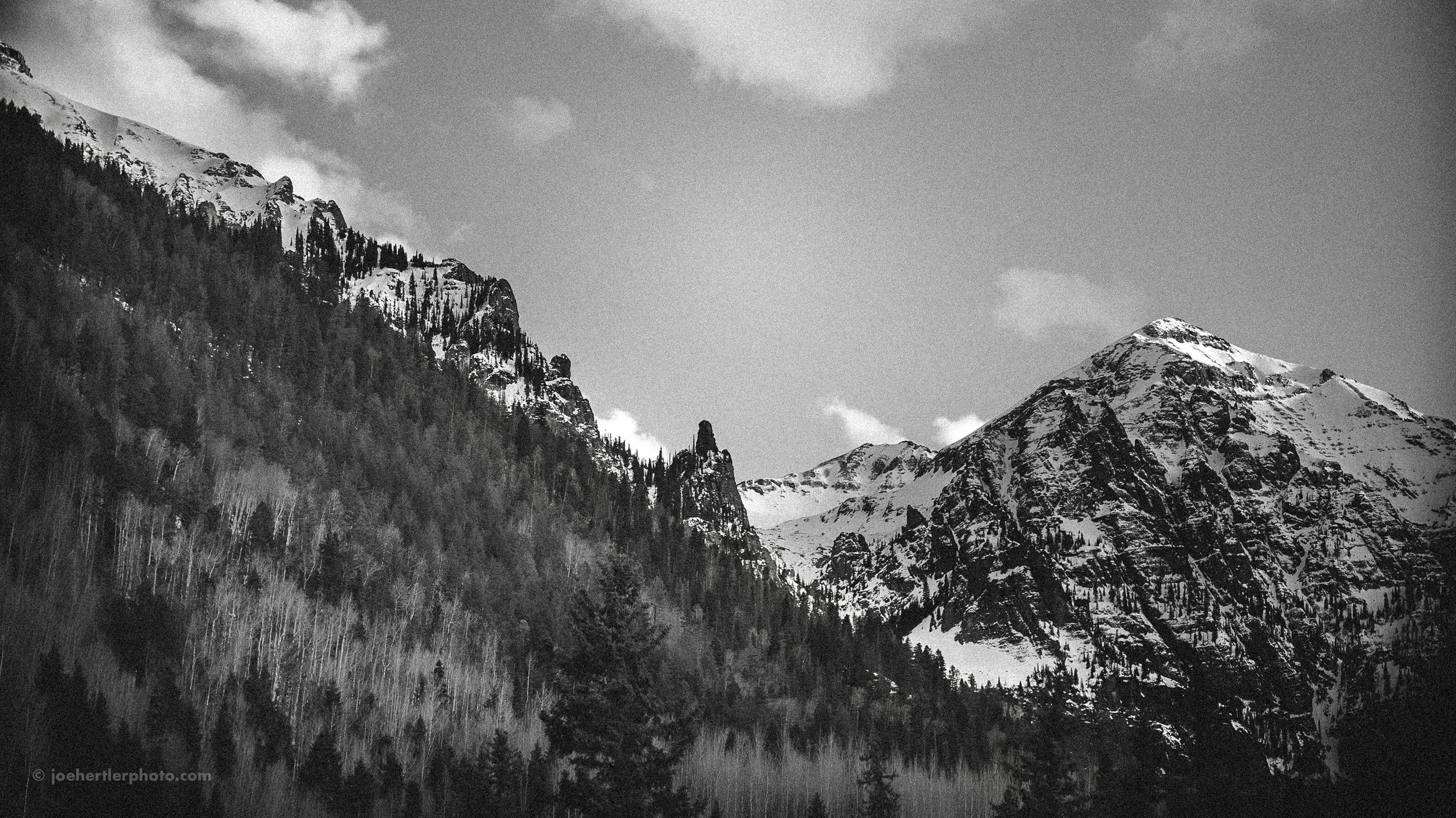 Black and white photo of snow-covered mountain peaks with a forest at the base.