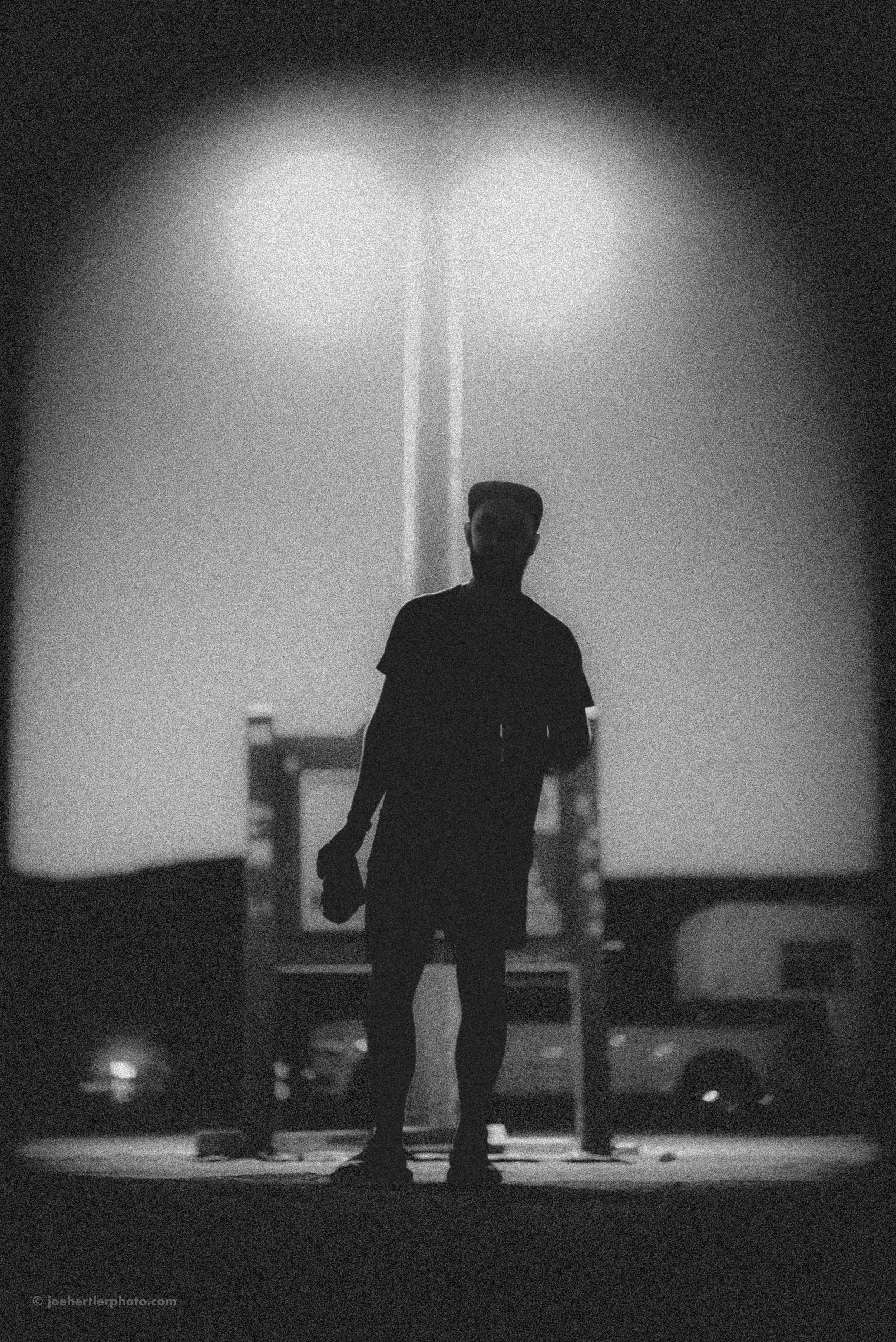 Silhouette of a man in black clothing standing outdoors at night, with a bright light source creating a halo effect in the background.