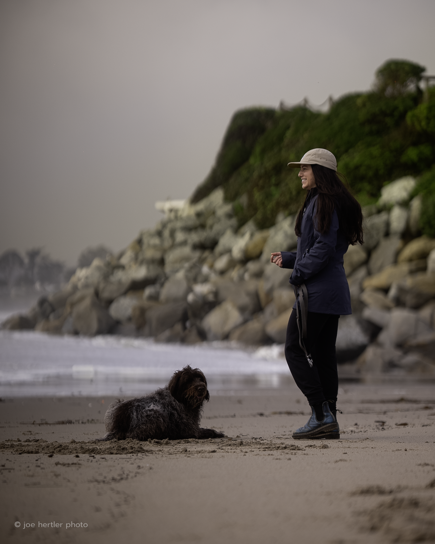 A woman and her dog on the beach, with rocky cliffs and greenery in the background, during overcast weather.