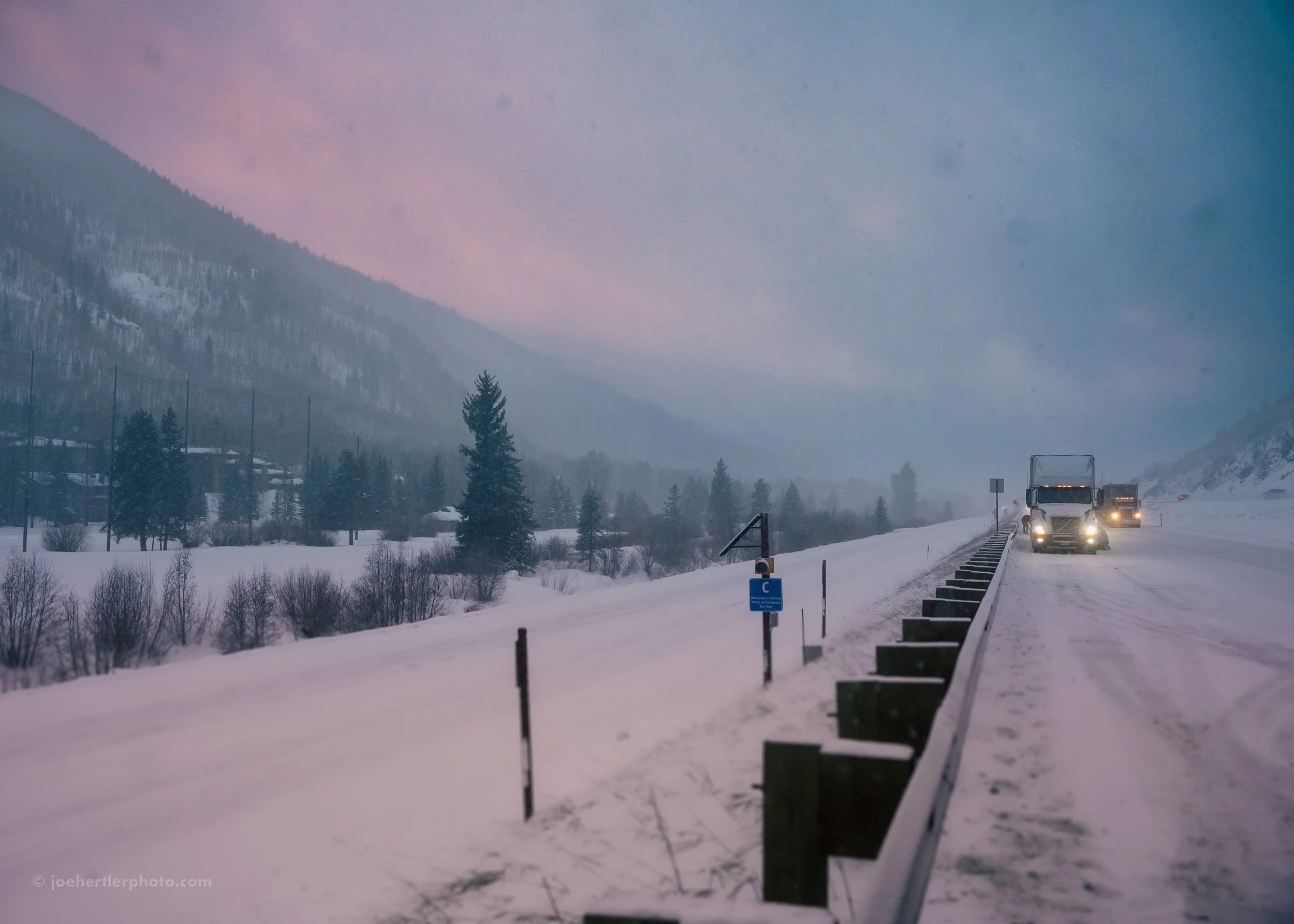 Snow-covered mountain landscape with a highway, several trucks driving, and trees in the distance at dusk or dawn.