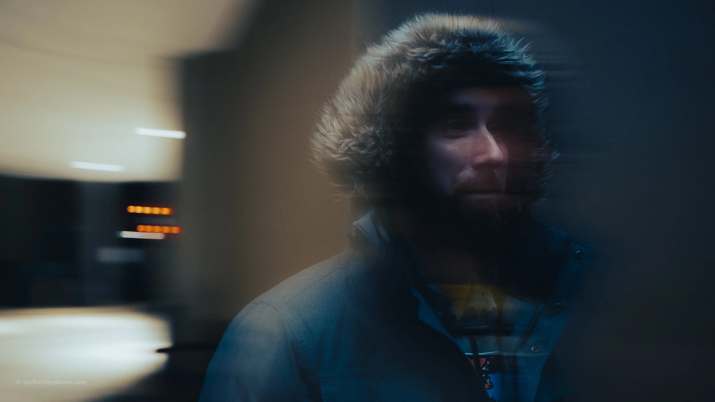 A person wearing a hooded winter jacket with a fur-lined hood, looking toward the camera, with blurred lights in the background.