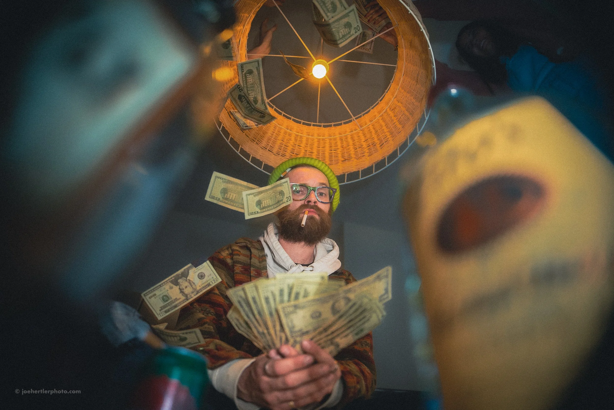 A man with a beard, glasses, and a yellow beanie smokes a cigarette and holds a fan of dollar bills. Dollar bills are also falling around him. The photo is taken from a low angle, showing a ceiling with a hanging light fixture and dollar bills attach