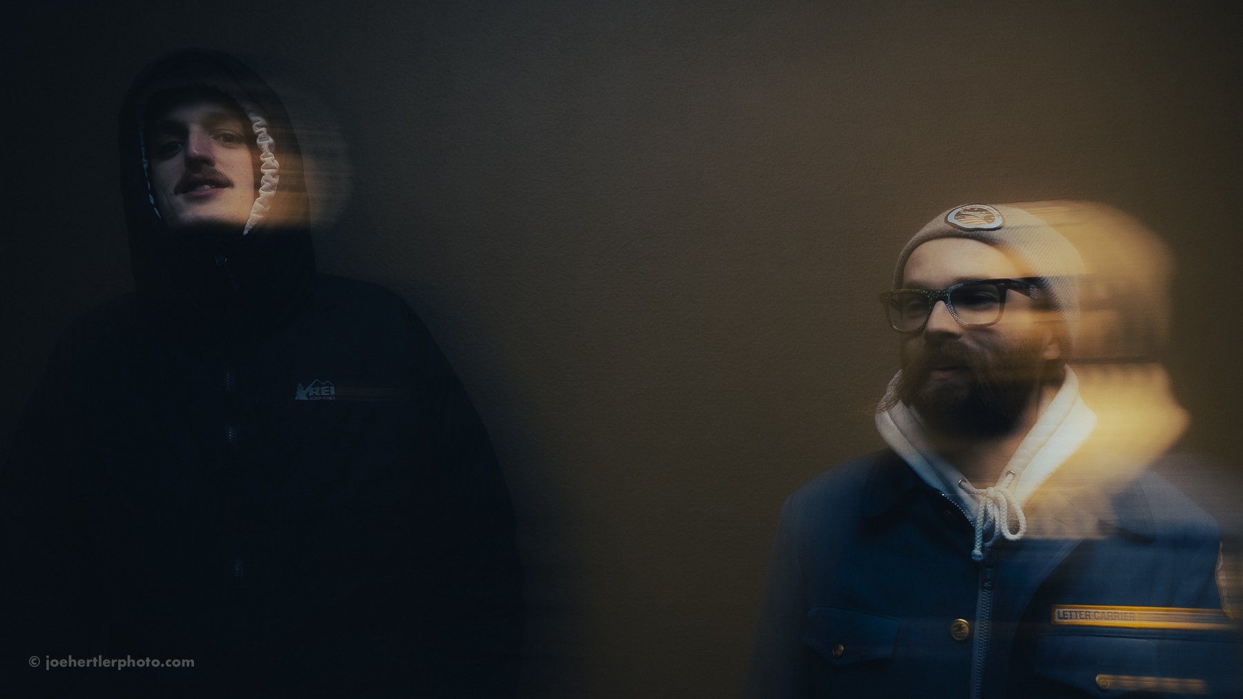 Two men standing against a dark background, one wearing a black hoodie with a reflective face and the other wearing glasses, a beanie, and a jacket with a patch that says "Letter Carrier".