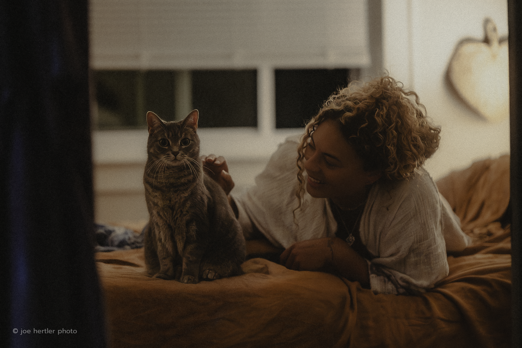 A woman with curly hair lying on a bed and smiling at a tabby cat sitting on the bed, inside a cozy room with soft lighting.