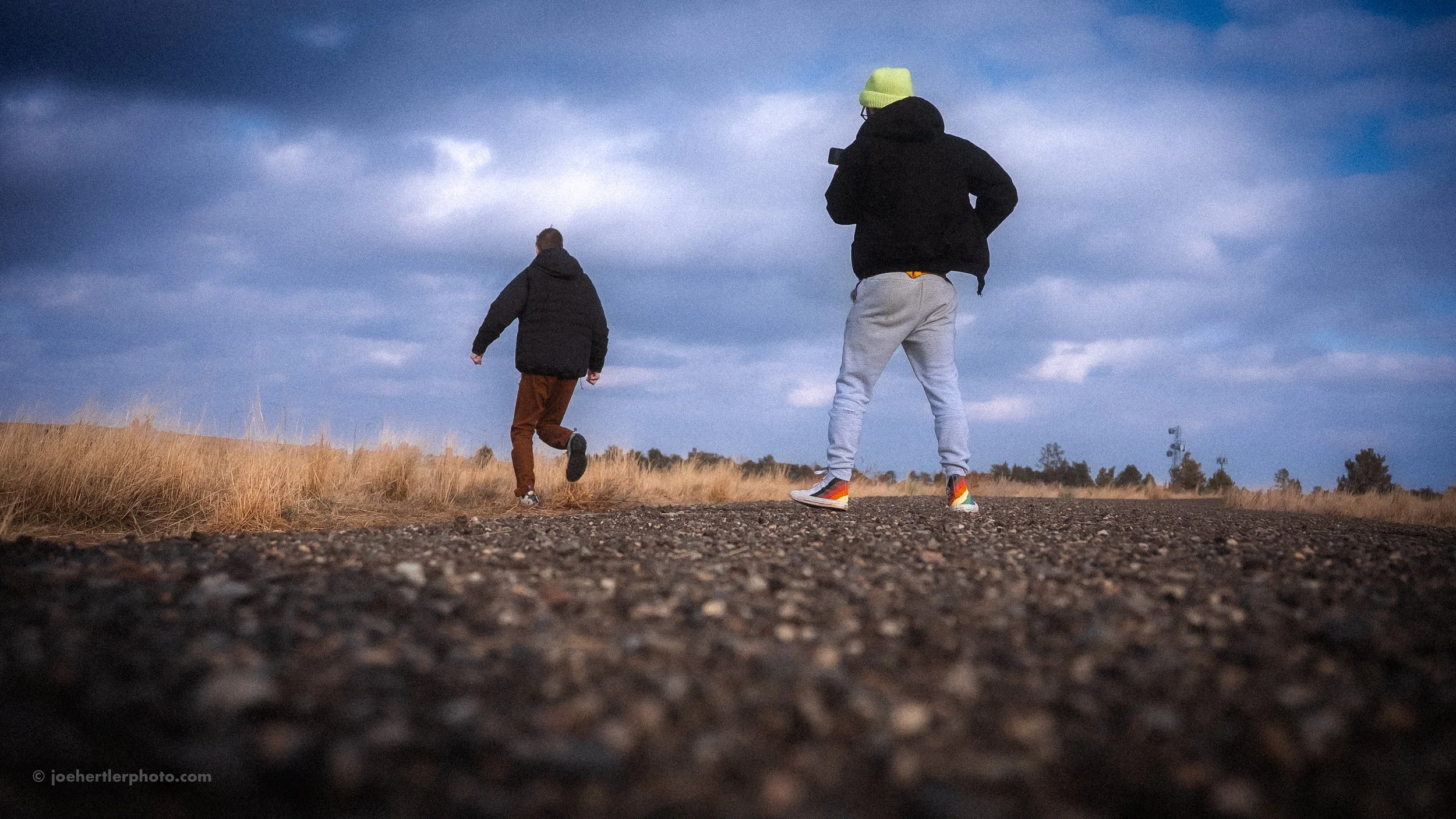 Two people walking on a gravel path in a field with tall grass and a cloudy sky overhead.