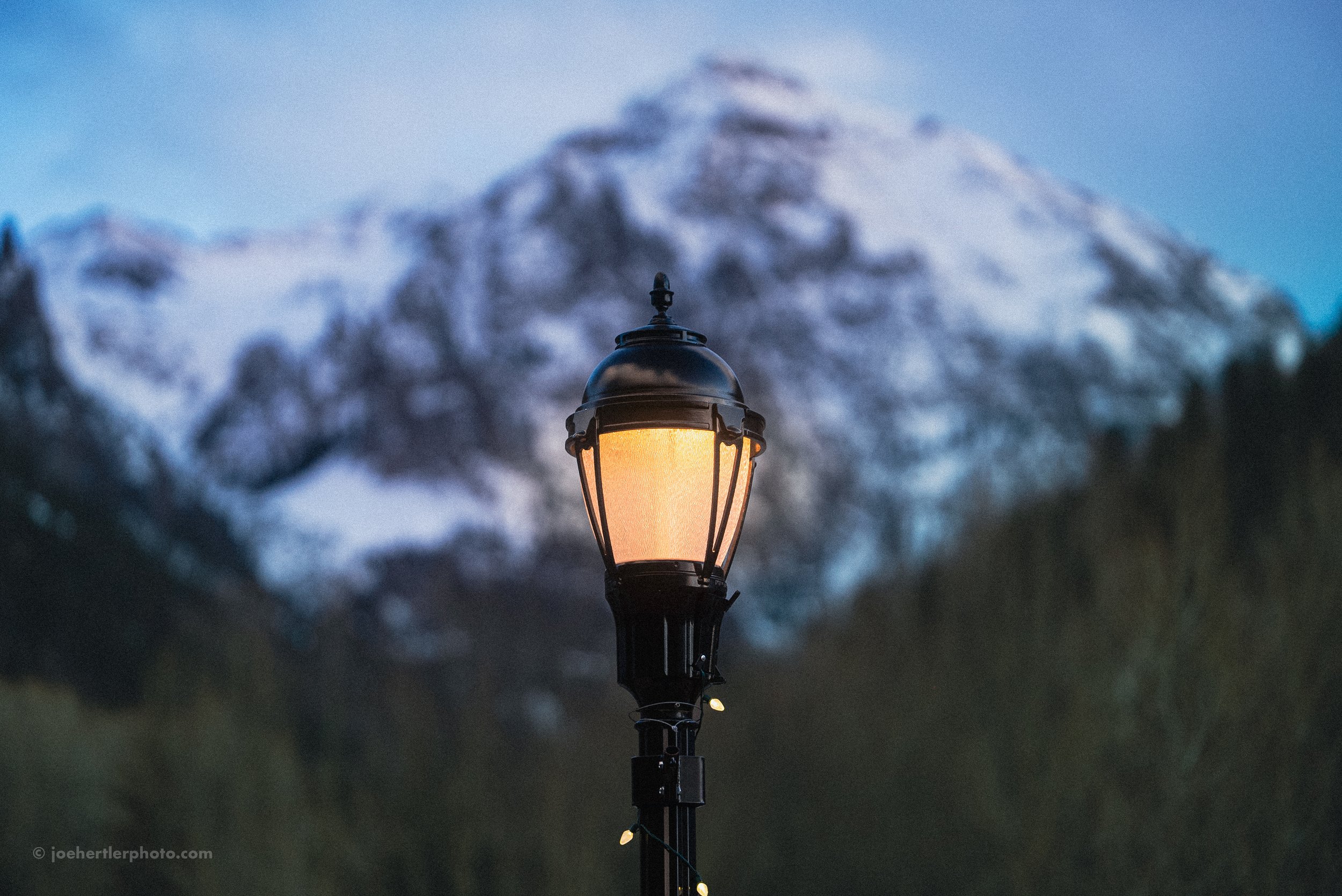 A street lamp with a glowing yellow light against a backdrop of snow-capped mountain and a partly cloudy evening sky.
