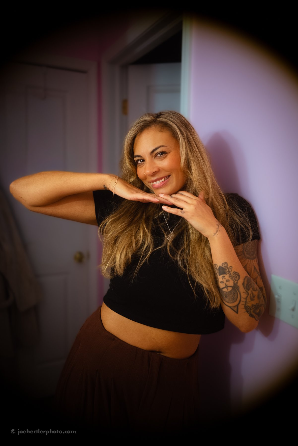 A young woman with long blonde hair, tattoos on her arm, wearing a black crop top and brown pants, smiling and posing indoors against a pink wall.