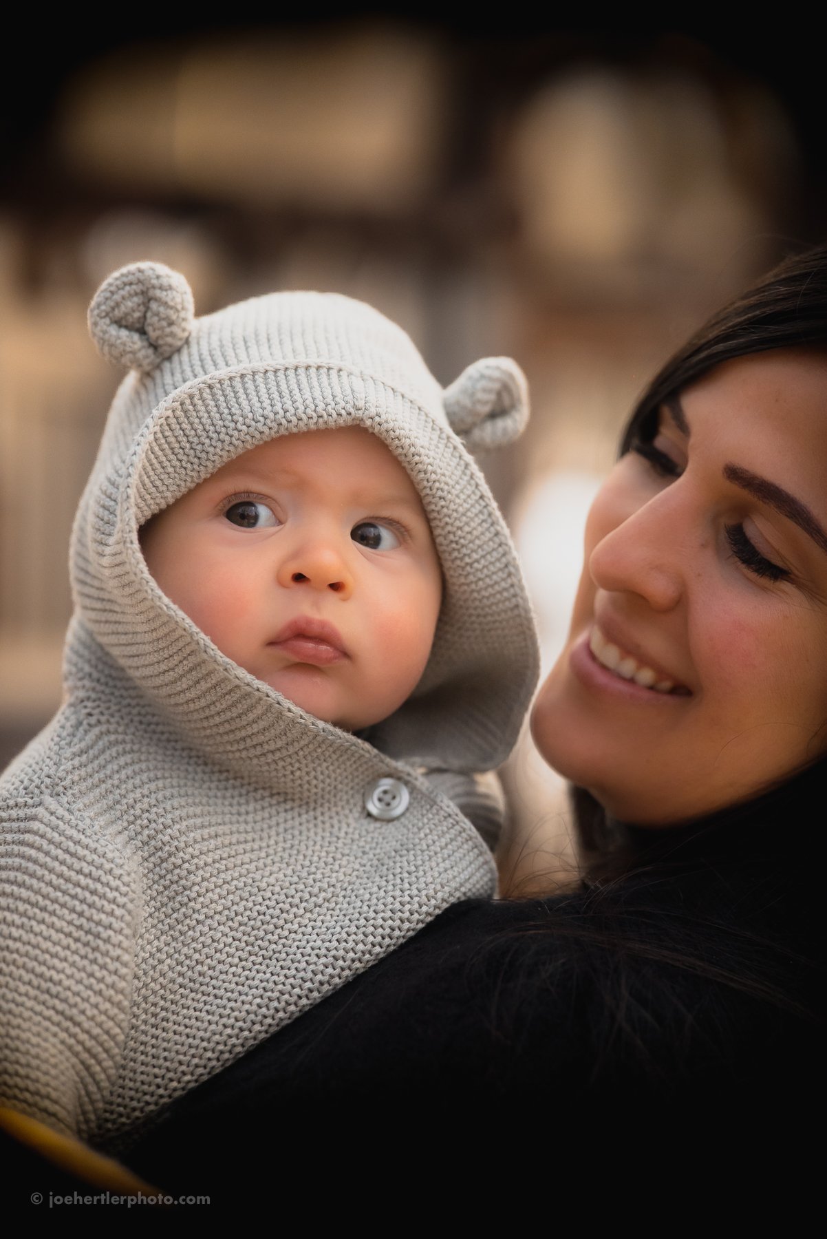 A smiling woman holding a baby dressed in a knit hoodie with bear ears, outdoors.
