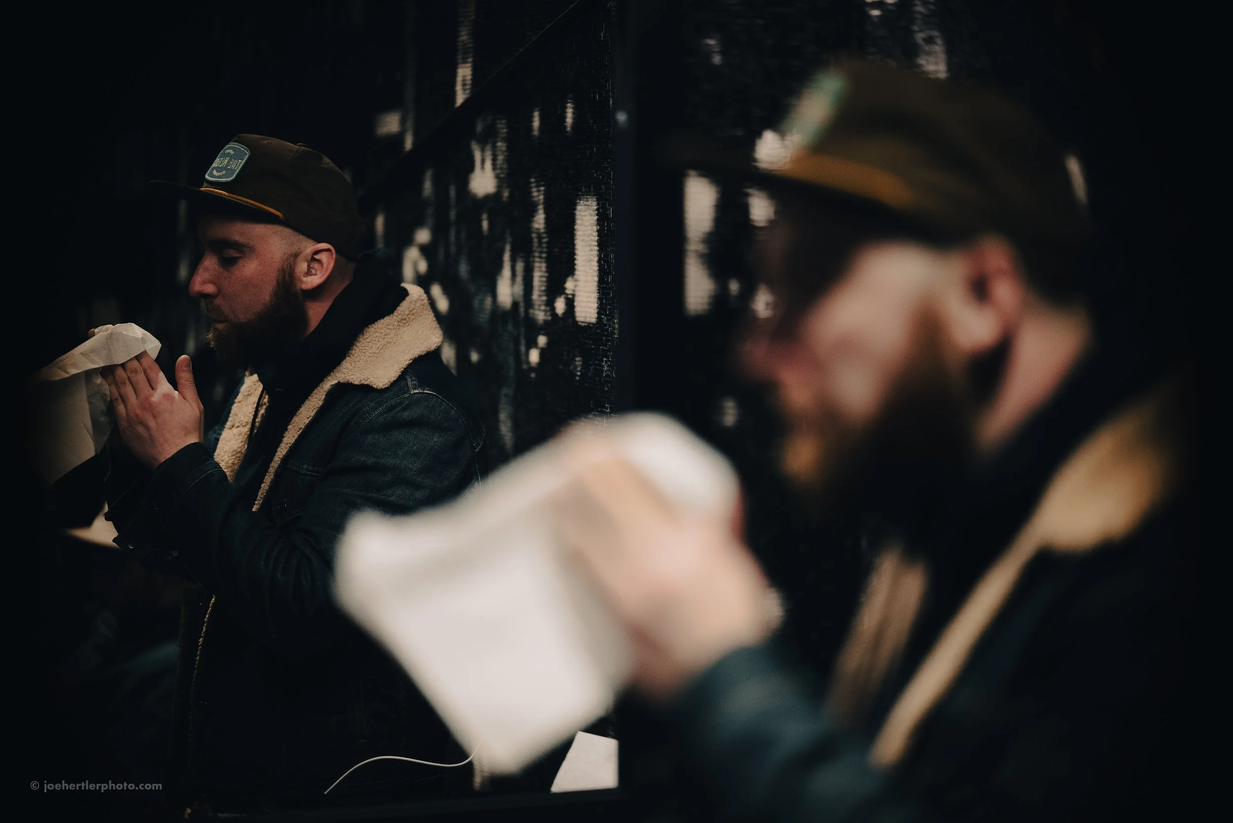 Two men sitting against a black wall, drinking from paper cups in a dark, intimate setting.