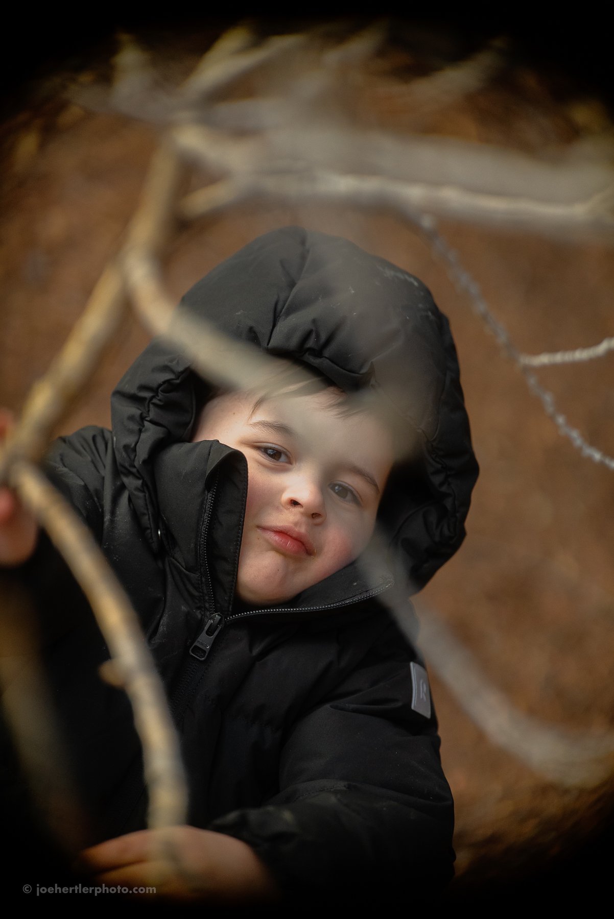 A young boy wearing a black hooded jacket looks through a hole in a chain-link fence.