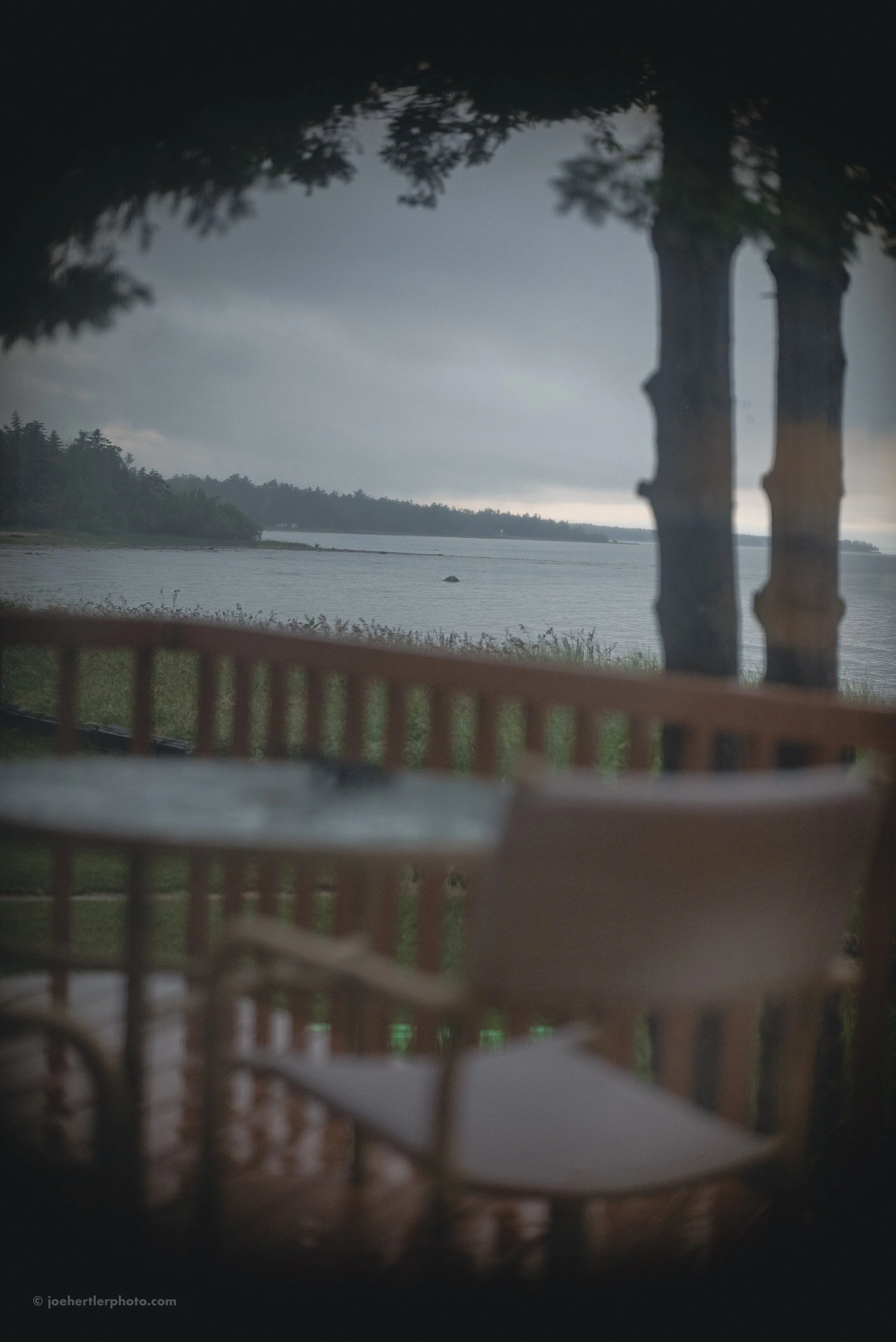 View of a lake through a window with a wooden bench outside. Dark clouds over the water, trees on the horizon.