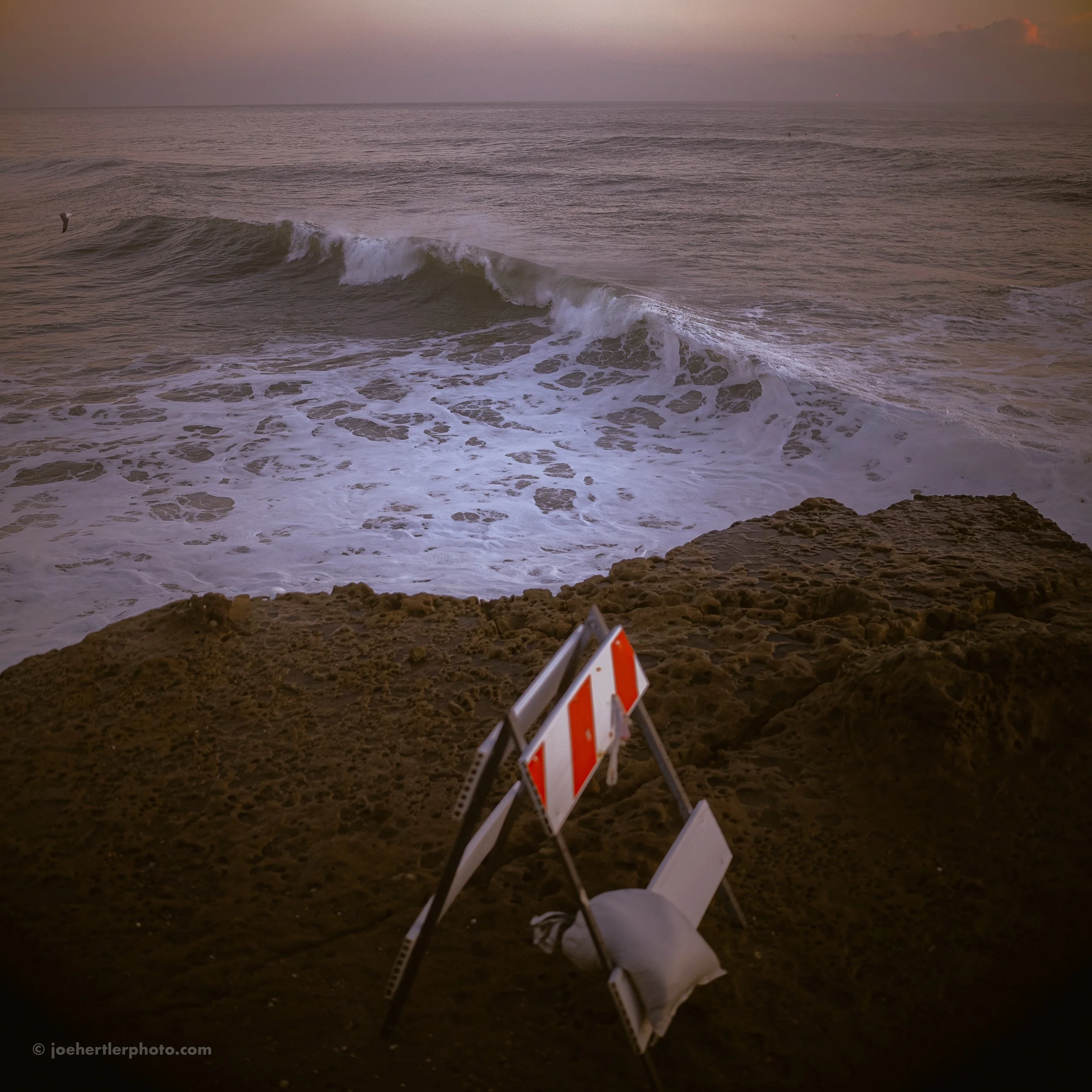 Ocean waves crashing on rocks at sunset with construction barriers in the foreground.