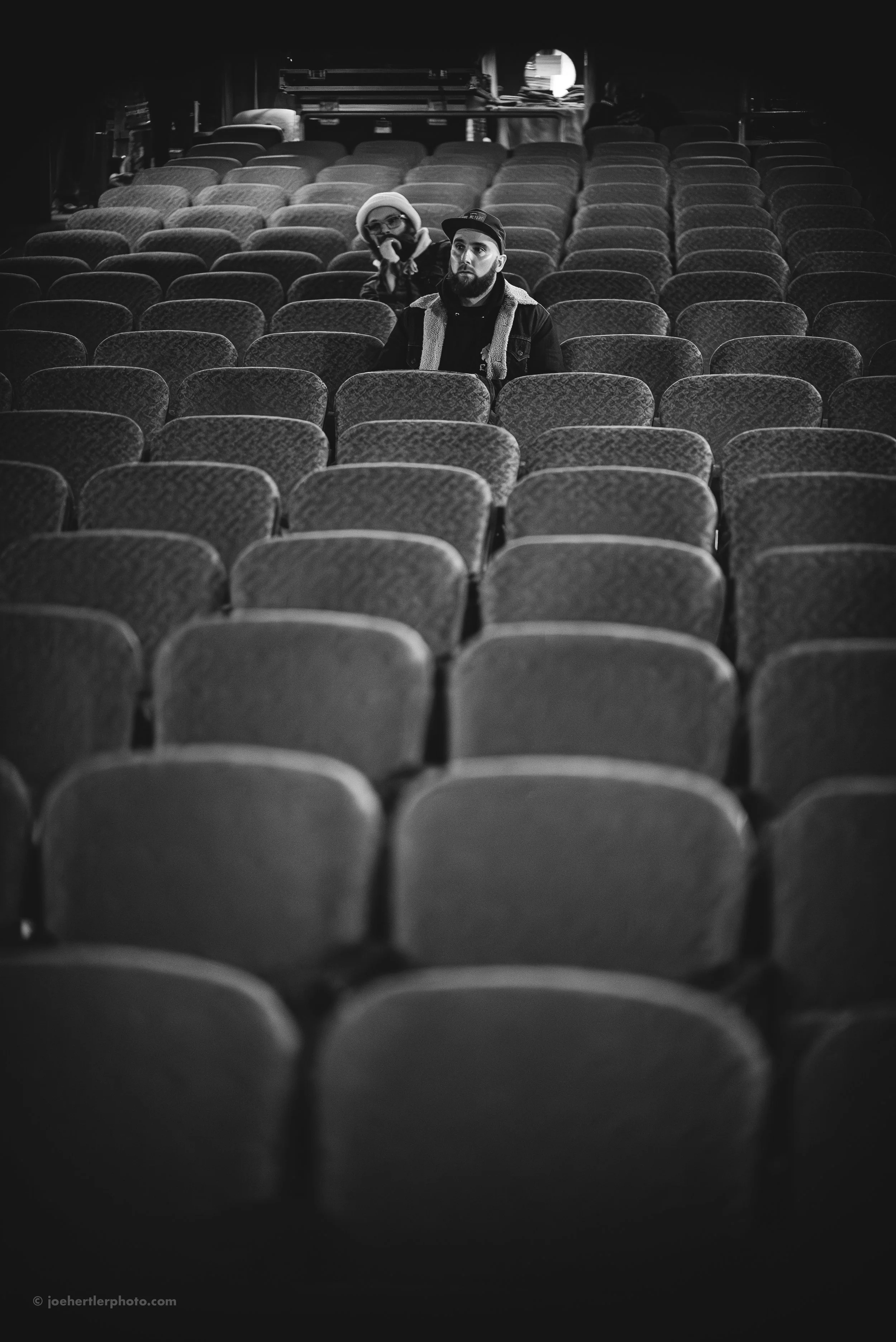 A black and white photo of an empty theater with rows of seats, with two people sitting in the middle rows. One person has a beard and is wearing a cap and a jacket, while the other person is wearing glasses and a hat, seen in the background.