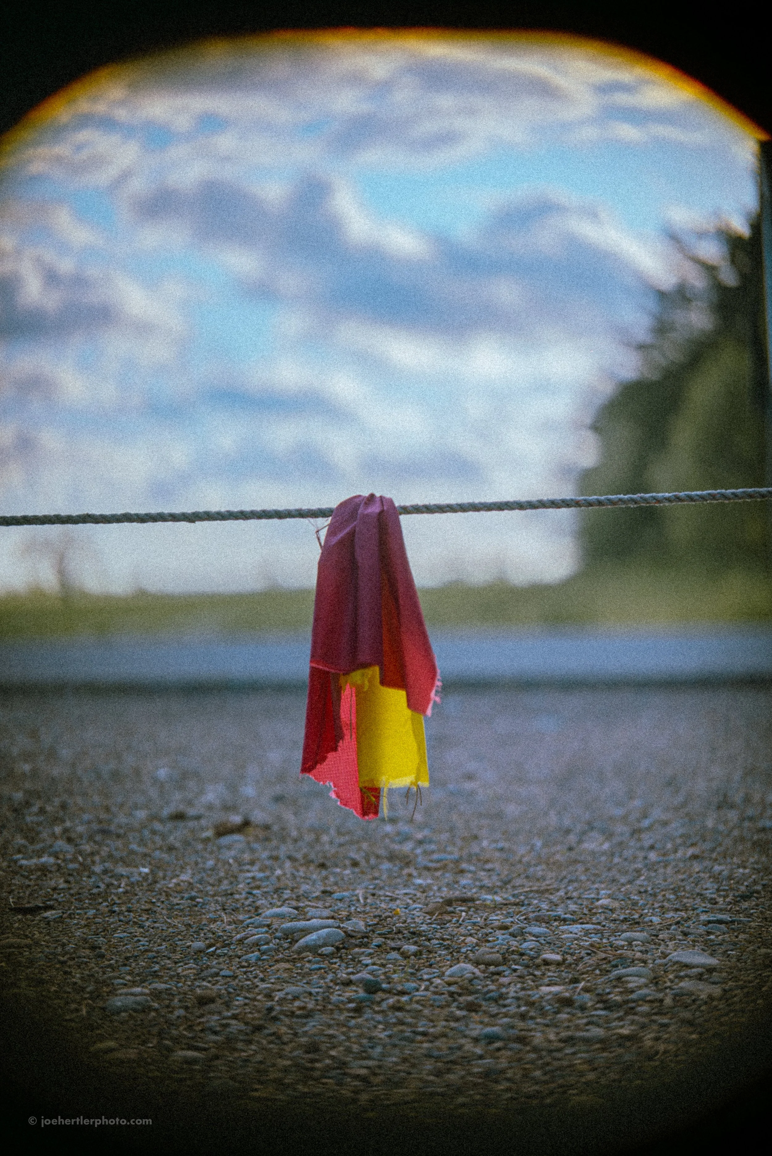 Colorful cloth hanging on a rope with a cloudy sky and distant trees in the background, viewed through a circular frame.