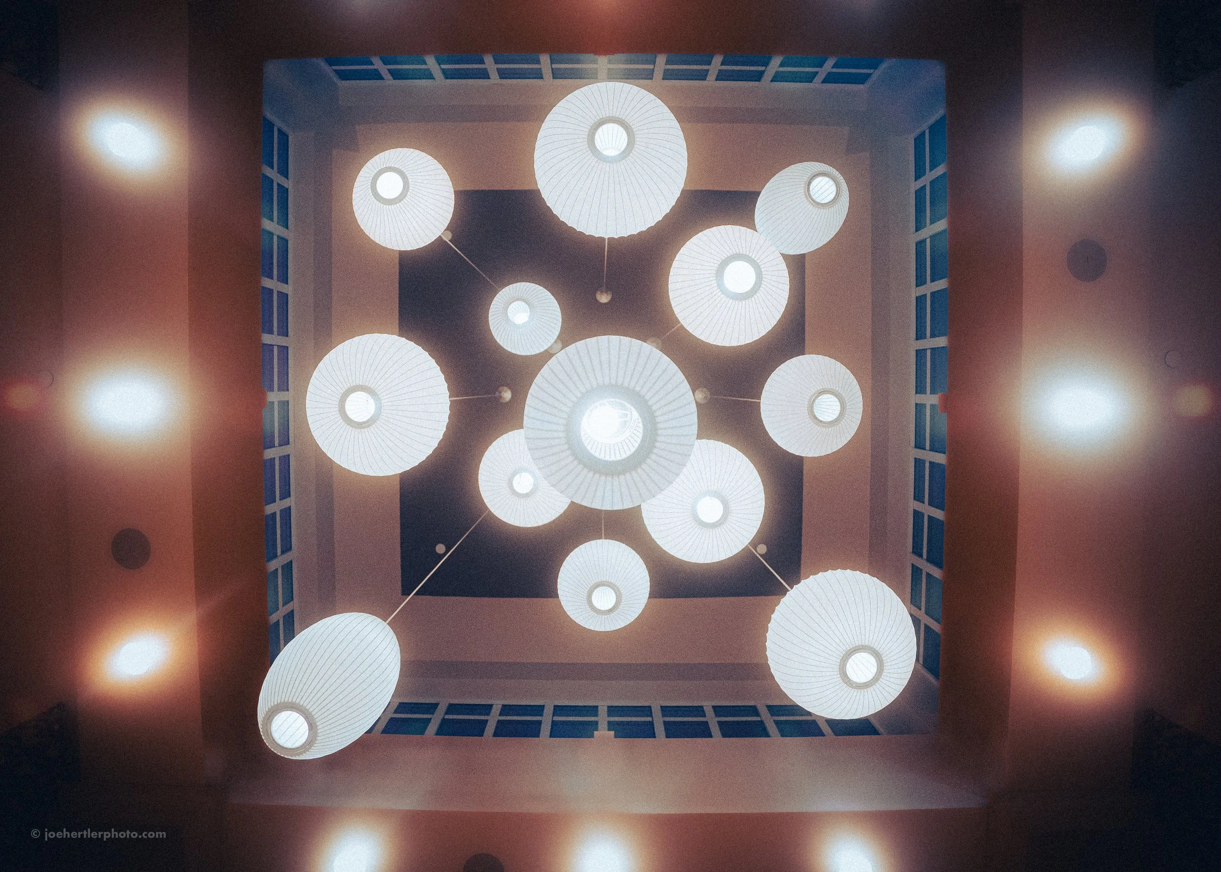 Ceiling view of multiple hanging pendant lights in a square room with blue window frames and warm wall lighting.