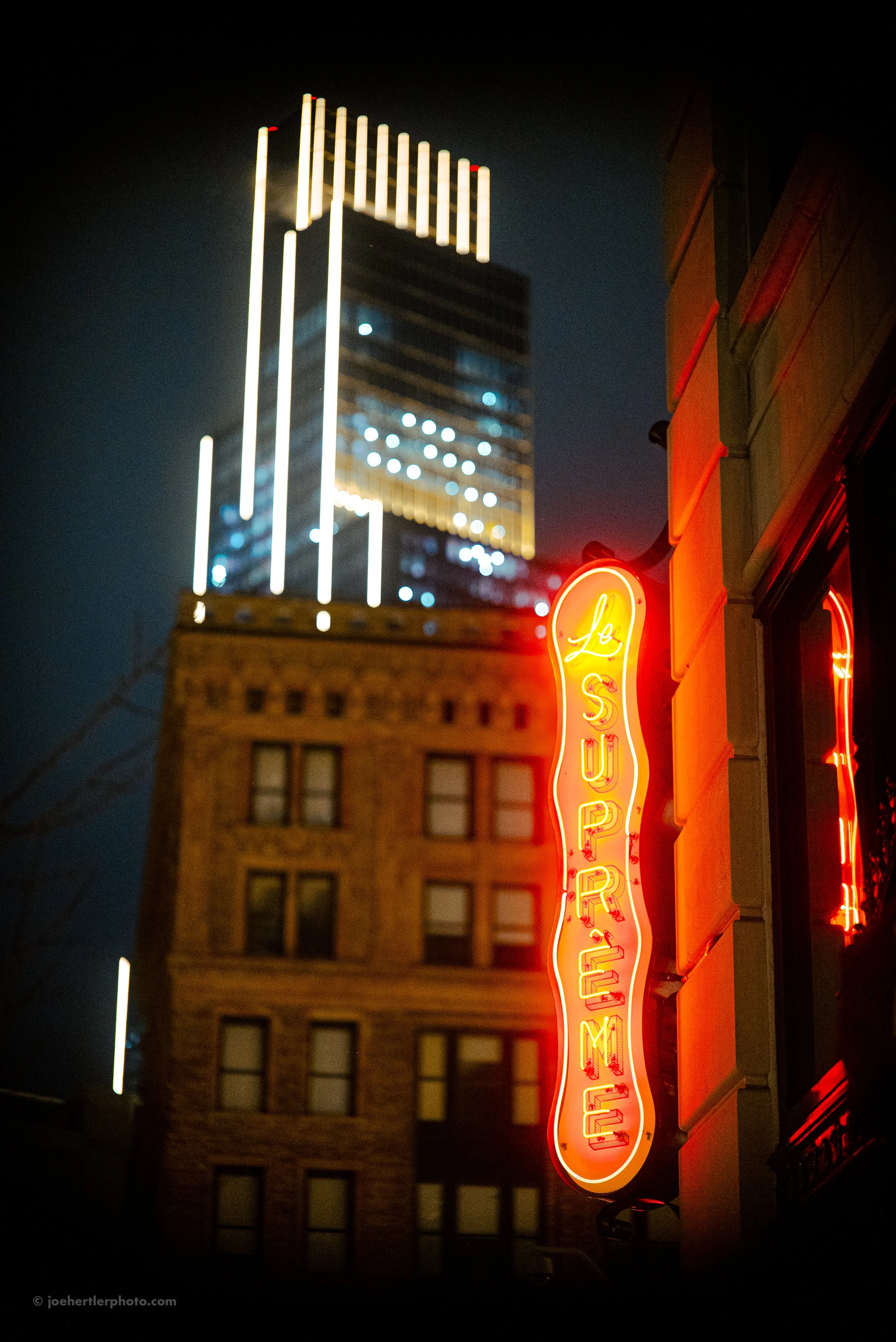 Night view of a city street with a red neon sign reading 'Le Suprem', a brick building, and a tall modern skyscraper with illuminated lines.
