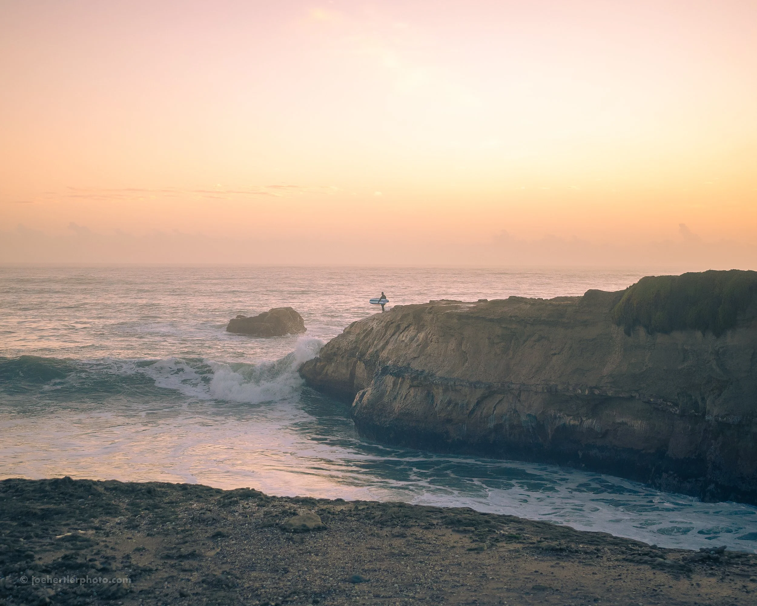 A person standing on a rocky cliff holding a surfboard at sunset, overlooking the ocean with waves crashing below.