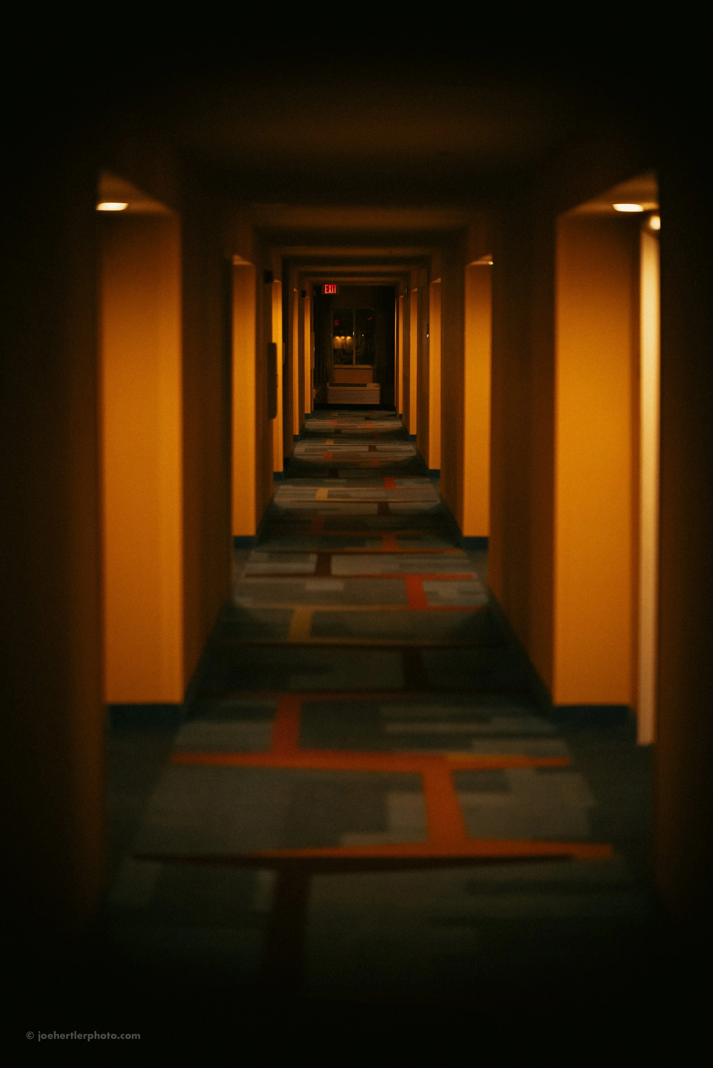 Dimly lit hotel corridor with yellow walls, patterned carpet, and exit sign at the end.