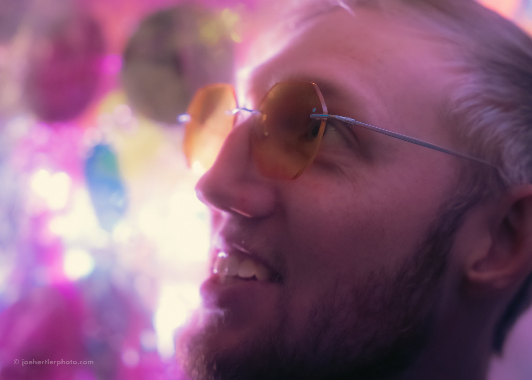 A man with blonde hair and beard wearing brown-tinted sunglasses, smiling in a colorful, vibrant setting with pink, purple, and blue lights.