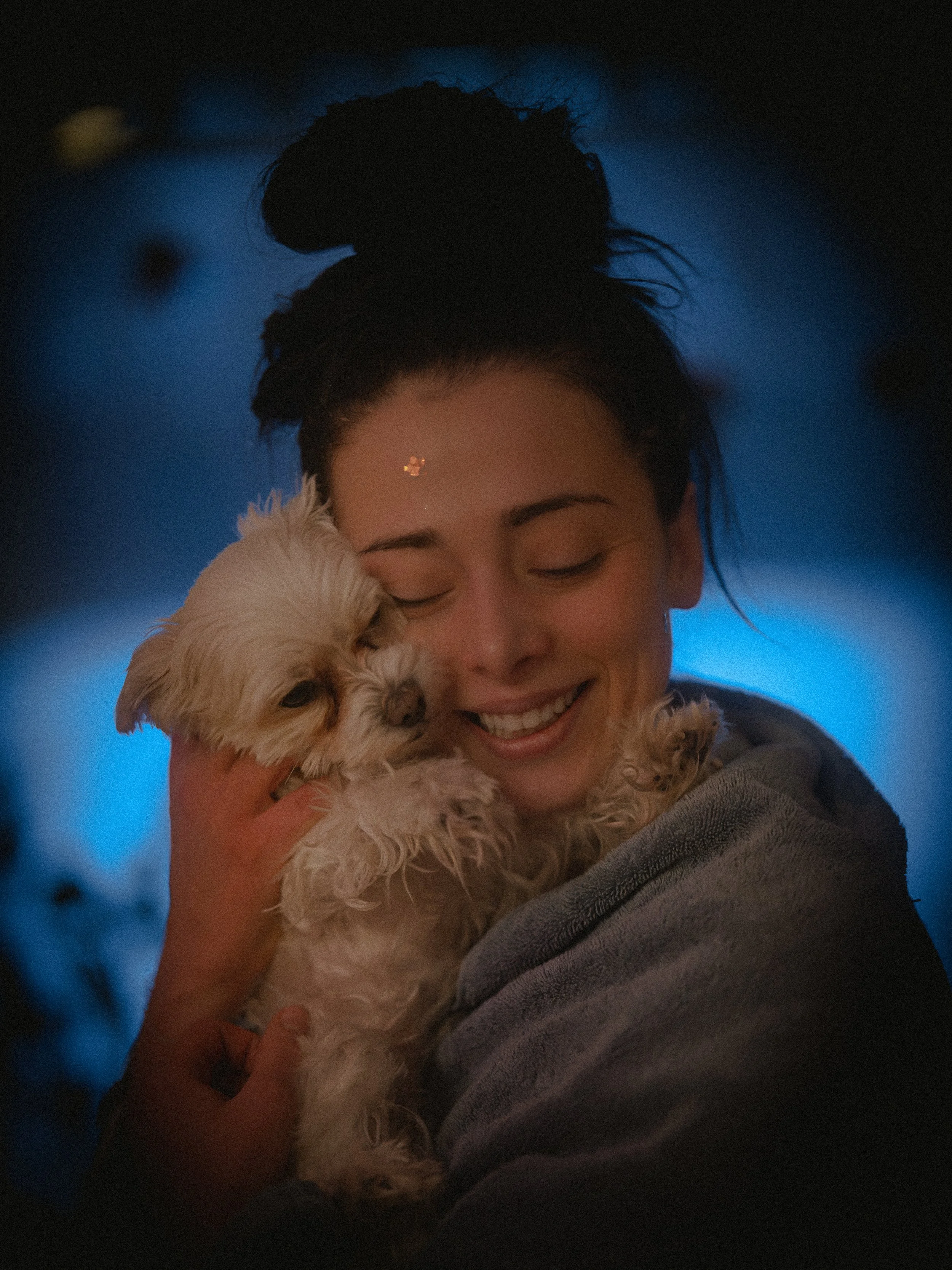 A woman with dark hair in a bun holding a small, fluffy, light-colored dog close to her face, smiling with her eyes closed, in a dimly lit setting with blue background lighting.