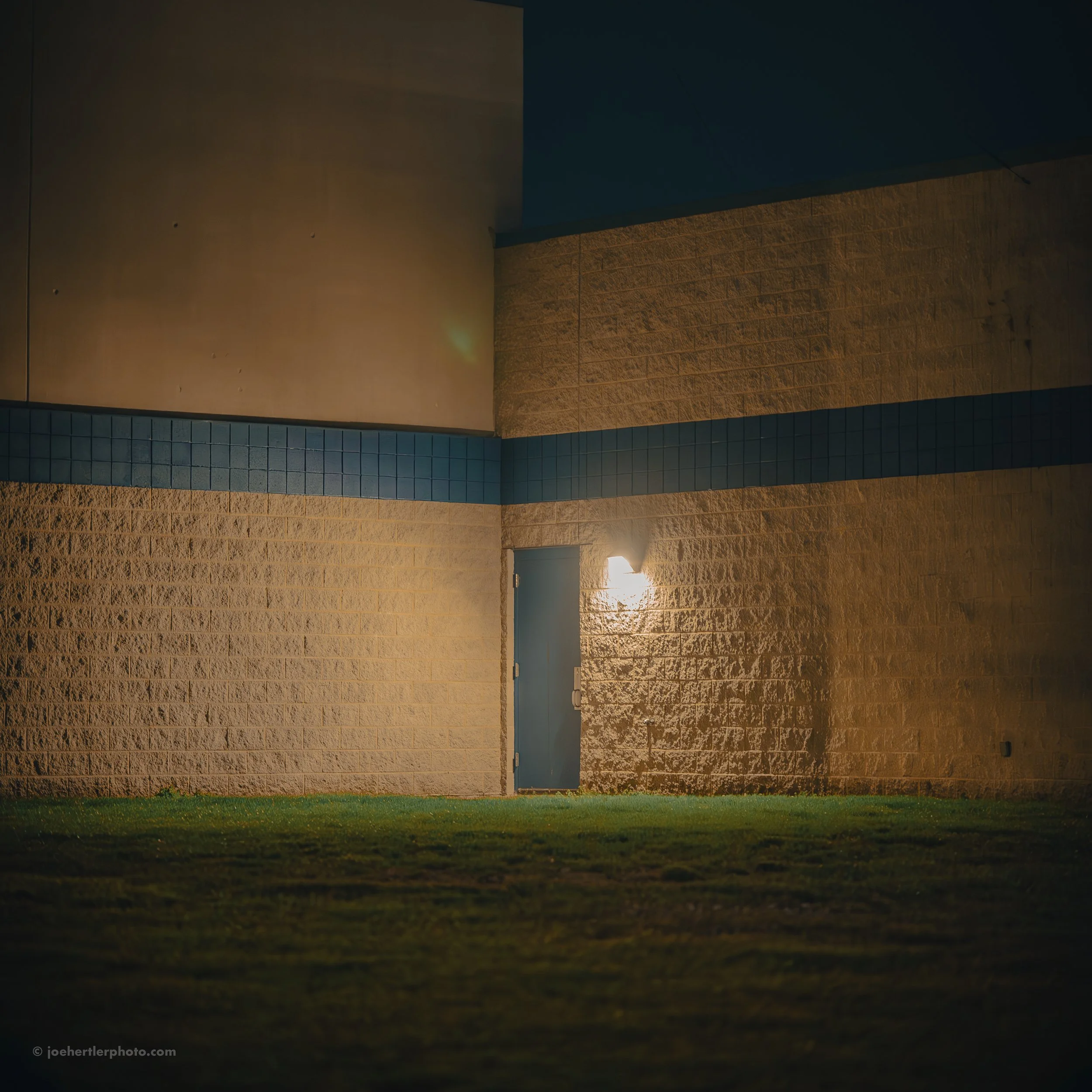 Nighttime exterior of a building with a blue door illuminated by a wall-mounted light, with a grassy foreground.