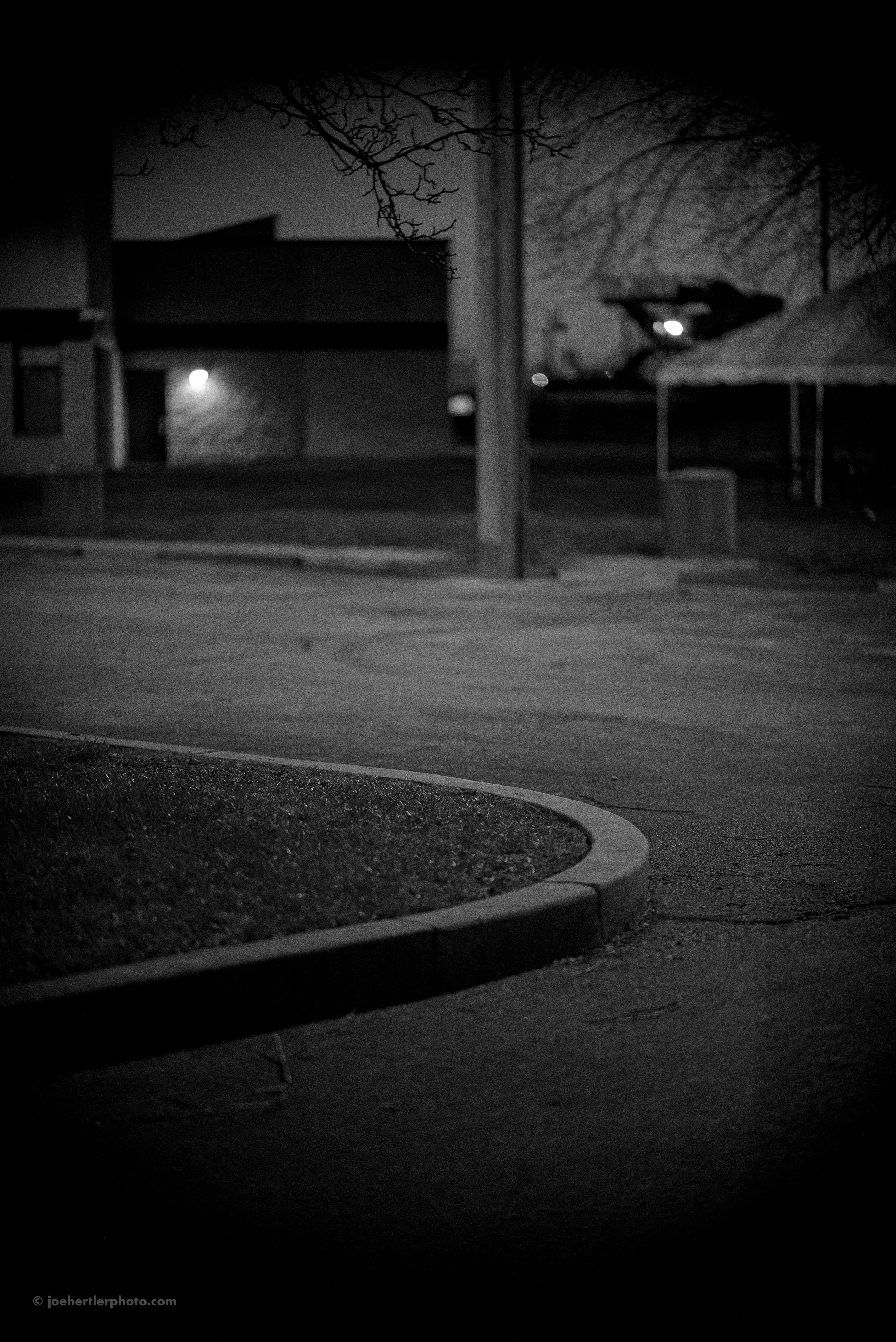 A black and white photo of a deserted parking lot at night, with a small patch of grass and a curved curb in the foreground, and a building, trees, and a pavilion with a roof in the background.