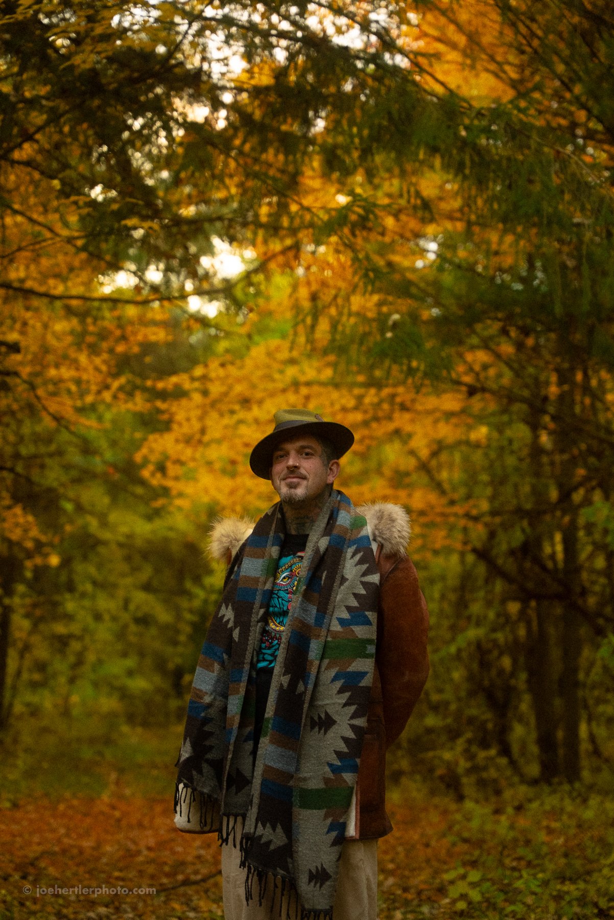 A man standing in a forest with autumn-colored leaves, wearing a hat, a bright scarf, and a coat with a fur hood.
